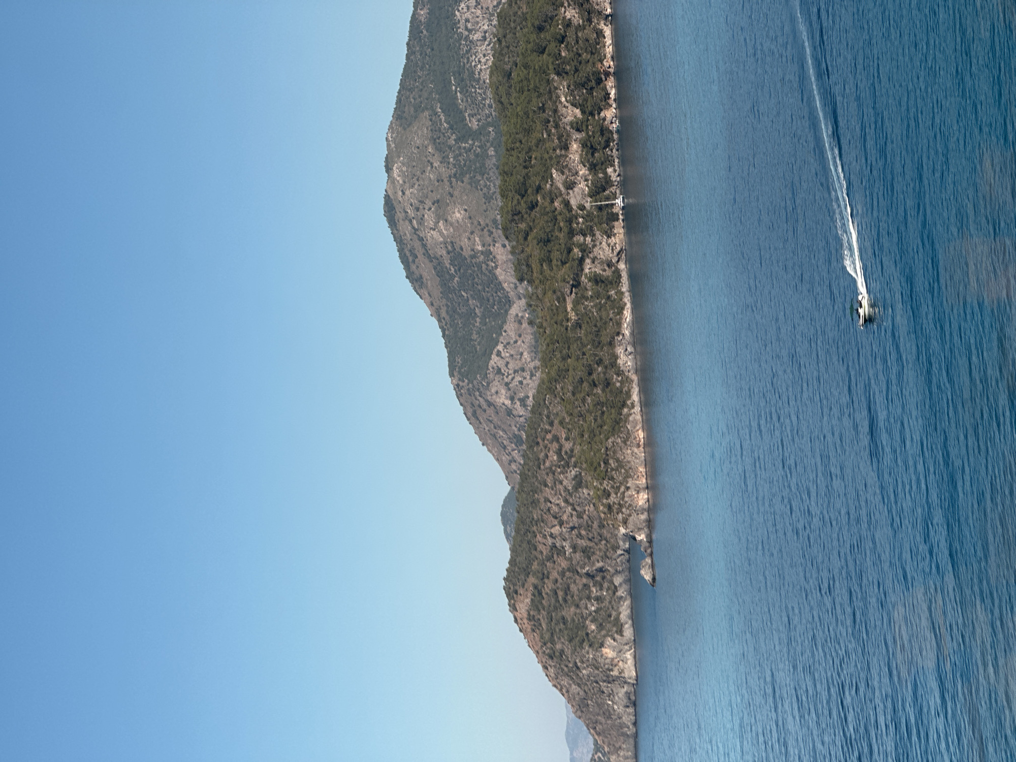 The scene is a coastal landscape with a mountain range bordering the sea. The mountains are covered in vegetation. The sea is blue and there is a small boat leaving a white trail behind it. It is likely located near Fethiye, Turkey based on the request.