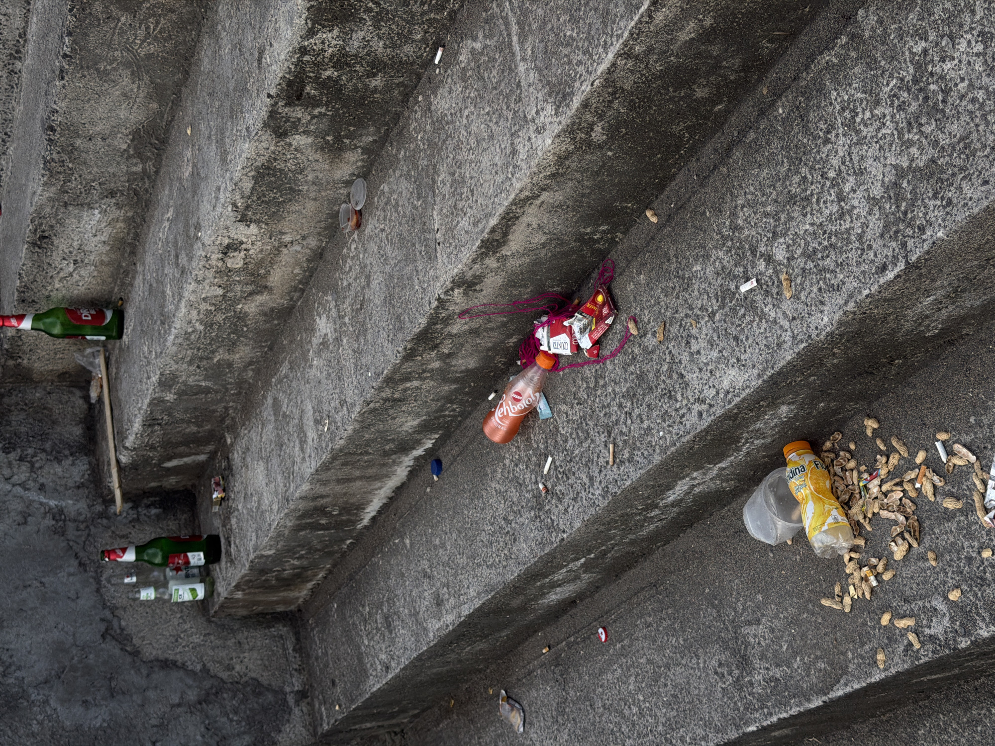 The media file captures a top-down view of a concrete staircase cluttered with various discarded items, creating a scene of urban litter. The steps are made of rough, dark grey concrete, showing signs of wear and possibly moss or dirt accumulation.
The primary subjects are numerous pieces of trash scattered across the stairs and in the recesses between steps. These include several green glass bottles, consistent with beer bottles, with the word "BINTANG" visible on at least two labels. There are also plastic bottles, such as an orange one clearly labeled "TEHBOTOL ORIGINAL," a popular Indonesian bottled tea, and a yellow one with "Pulpy" and "MINUMAN ... BUAH" (likely "Minuman Sari Buah" or fruit juice) visible.
Other discarded items include a large quantity of peanut shells, especially on one of the wider steps, numerous cigarette butts, clear plastic cups, small plastic containers with lids, and various plastic wrappers and pieces of debris. A red and white piece of packaging with the word "COUNTRY" is also visible, entangled with some red yarn or string.
No people are visible in the image, and there is no specific activity or event taking place other than the aftermath of items being discarded. The scene conveys a sense of neglect and untidiness. The lighting suggests it is daytime. Given the location context of North Kuta, Indonesia, the presence of specific Indonesian brand names like Bintang beer and Tehbotol reinforces the geographical setting. The image is a stark depiction of public litter on a set of concrete steps.