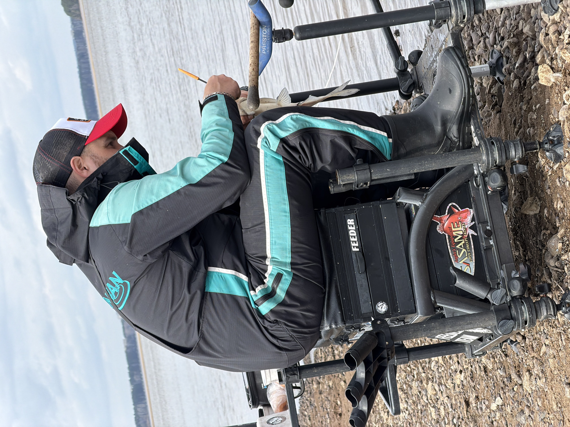 A man is seated in a black fishing platform on a rocky shoreline near a body of water, possibly the Kama Reservoir, near Chaykovsky, Russia. He wears a black and teal jacket, black pants with a teal stripe, and black boots. A red and white baseball cap covers his head. The fishing platform is equipped with accessories like a "FEEDER" box and a "SASAME" sticker. He is handling a small fish. A fishing rod is propped nearby. The sky is overcast.
