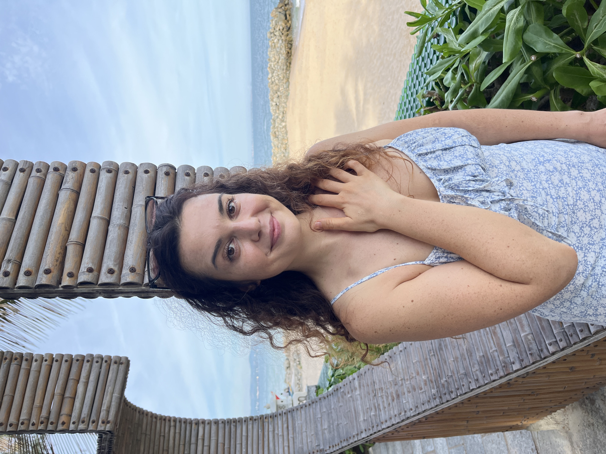 A woman with curly brown hair stands on a balcony overlooking a beach in Mũi Né, Vietnam. She is wearing a light blue floral dress and has her hand on her chest. Sunglasses are perched on her head. The background shows a sandy beach, the ocean, and a rocky breakwater. The balcony is constructed of bamboo.