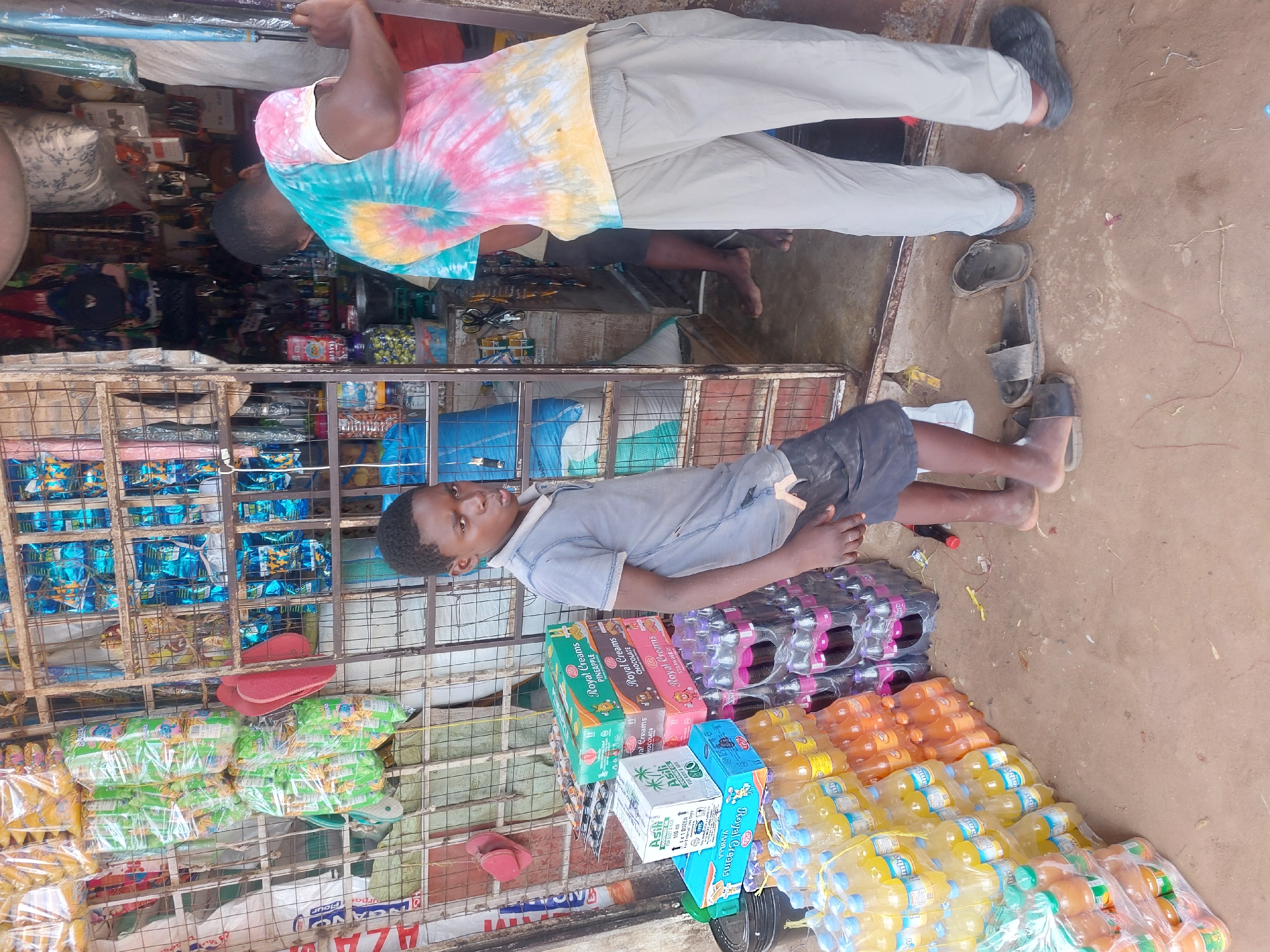 Here is a description of the image:
**Scene:** The image shows a bustling street scene likely in Dar es-Salaam, Tanzania. A small shop is visible with a variety of goods on display.
**People:** There are two people visible. One man is wearing a colorful tie-dye shirt and light-colored pants, standing in front of the shop. A younger boy is wearing a grey shirt and dark shorts, appearing to be near the items for sale.
**Objects:** The shop contains a wide array of items including:
* Bottled drinks of various colors and brands (likely juices and sodas)
* Packaged snacks (cookies and chips)
* Boxes of what appear to be food products (such as "Royal Creams")
* Other miscellaneous items inside the shop behind a metal grid.
**Location:** Based on the goods sold and the overall appearance of the shop and surroundings, it is very likely this is a street-side shop in Dar es-Salaam, Tanzania.