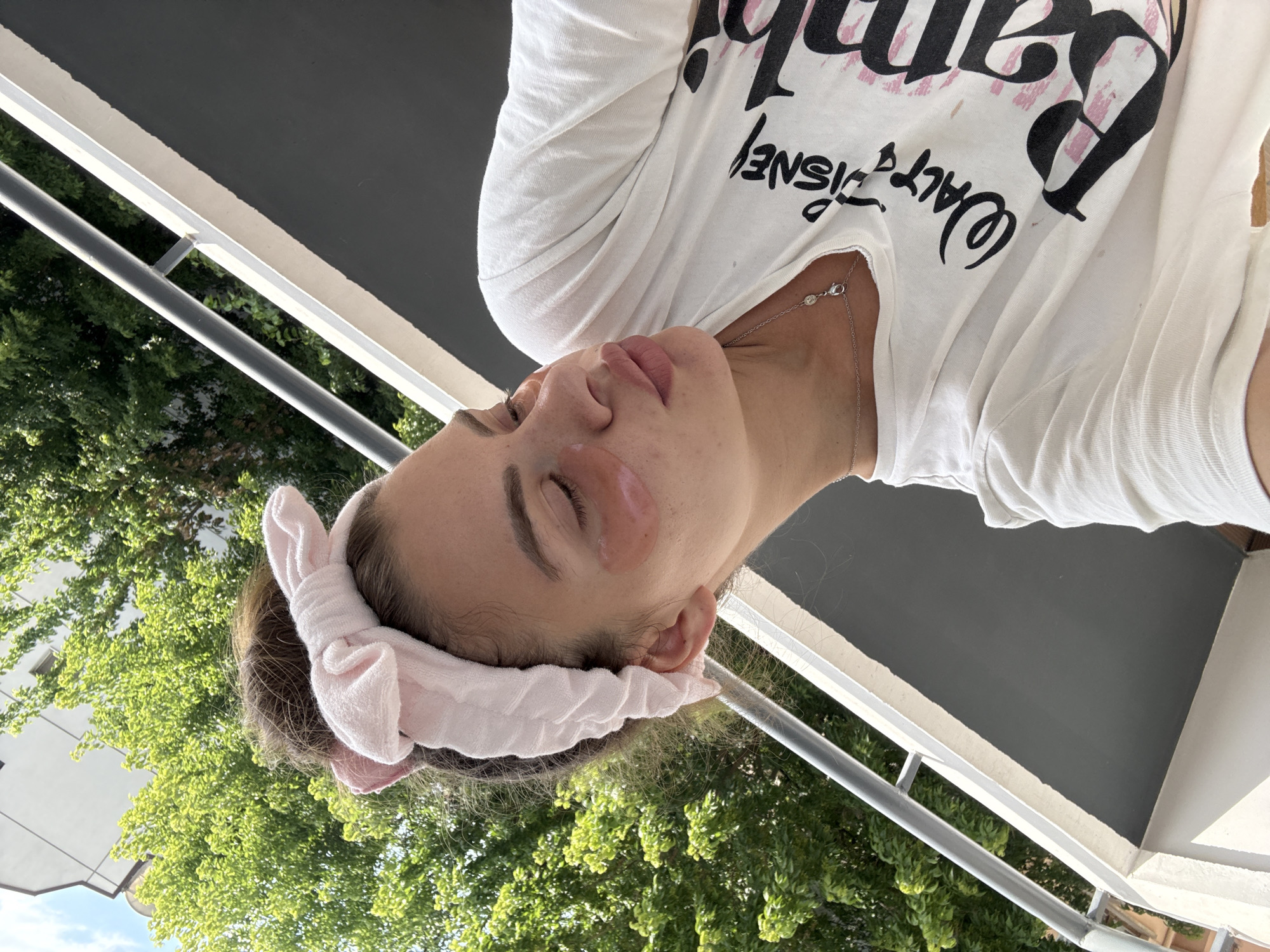 A young woman with brown hair pulled up and a pink headband is shown from the chest up, taking a selfie on a balcony. She is wearing a white t-shirt with a Walt Disney Bambi graphic on it, and a delicate silver necklace. She has pink under-eye patches on. The background shows a railing and lush green trees, with a white building visible through the leaves. The lighting suggests it is daytime. The location is a balcony in the city of Plovdiv, Bulgaria.