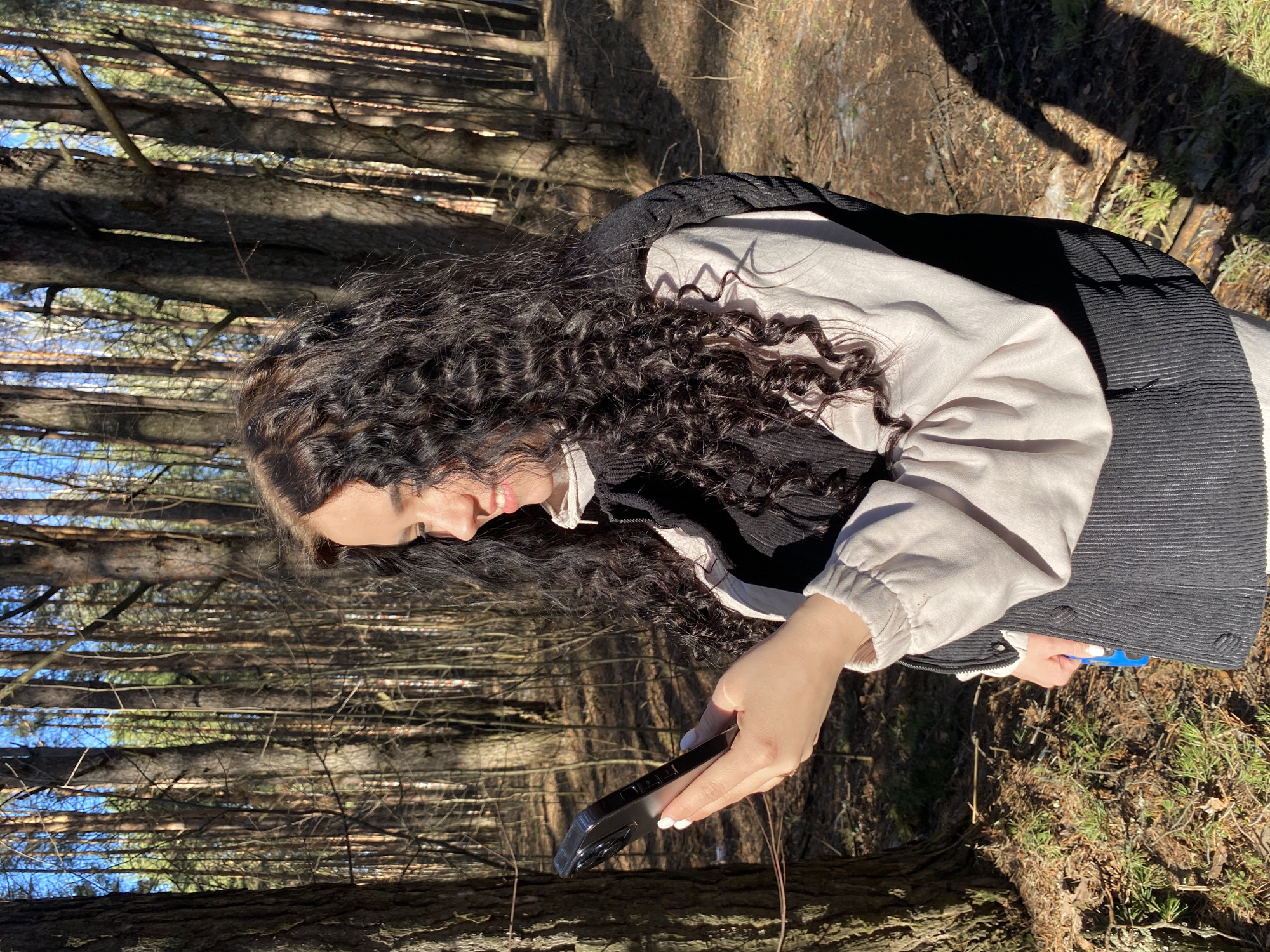 A woman with long, dark, curly hair is outside, looking down at a black smartphone in her hands. She is wearing a layered outfit consisting of a white shirt, a light-colored jacket with gathered sleeves, and a black vest. The background suggests a wooded area with many tall, thin trees and dappled sunlight on the ground. The location is Chaikovsky, Russia.