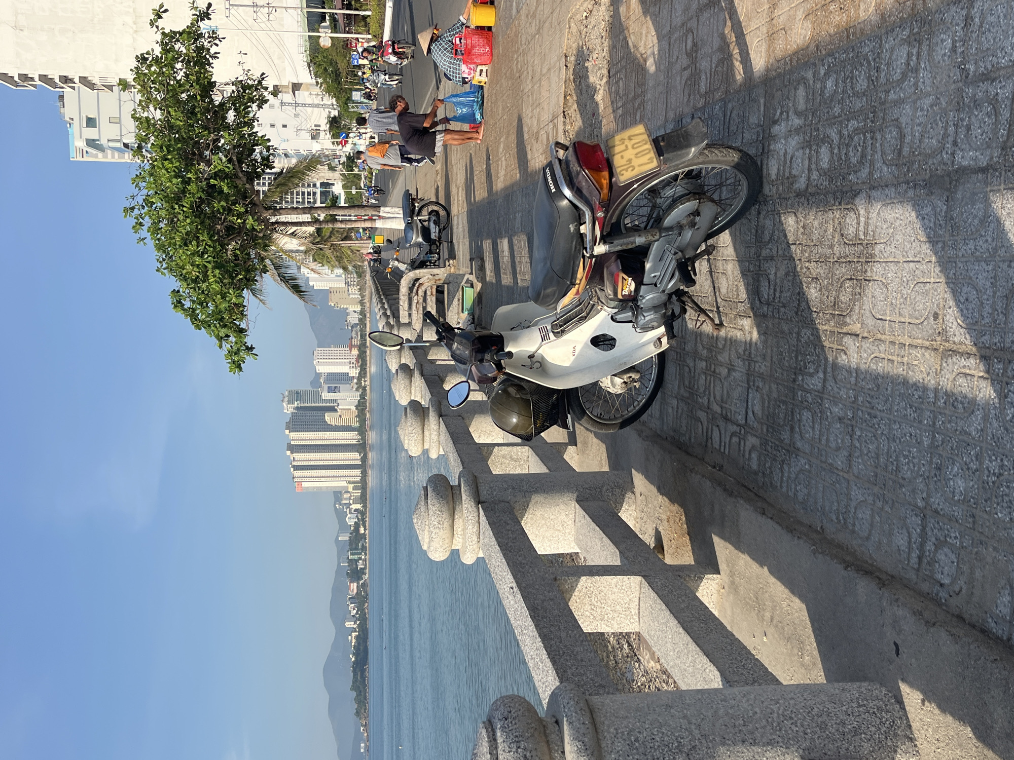 The image shows a sunny day scene on the waterfront in Bac Nha Trang Ward, Vietnam. A row of parked motorbikes lines a sidewalk paved with patterned gray tiles. A low, ornate concrete barrier separates the sidewalk from the water. Buildings rise in the distance across the bay. Several people are visible along the sidewalk, including a vendor wearing a conical hat. The sky is clear blue.