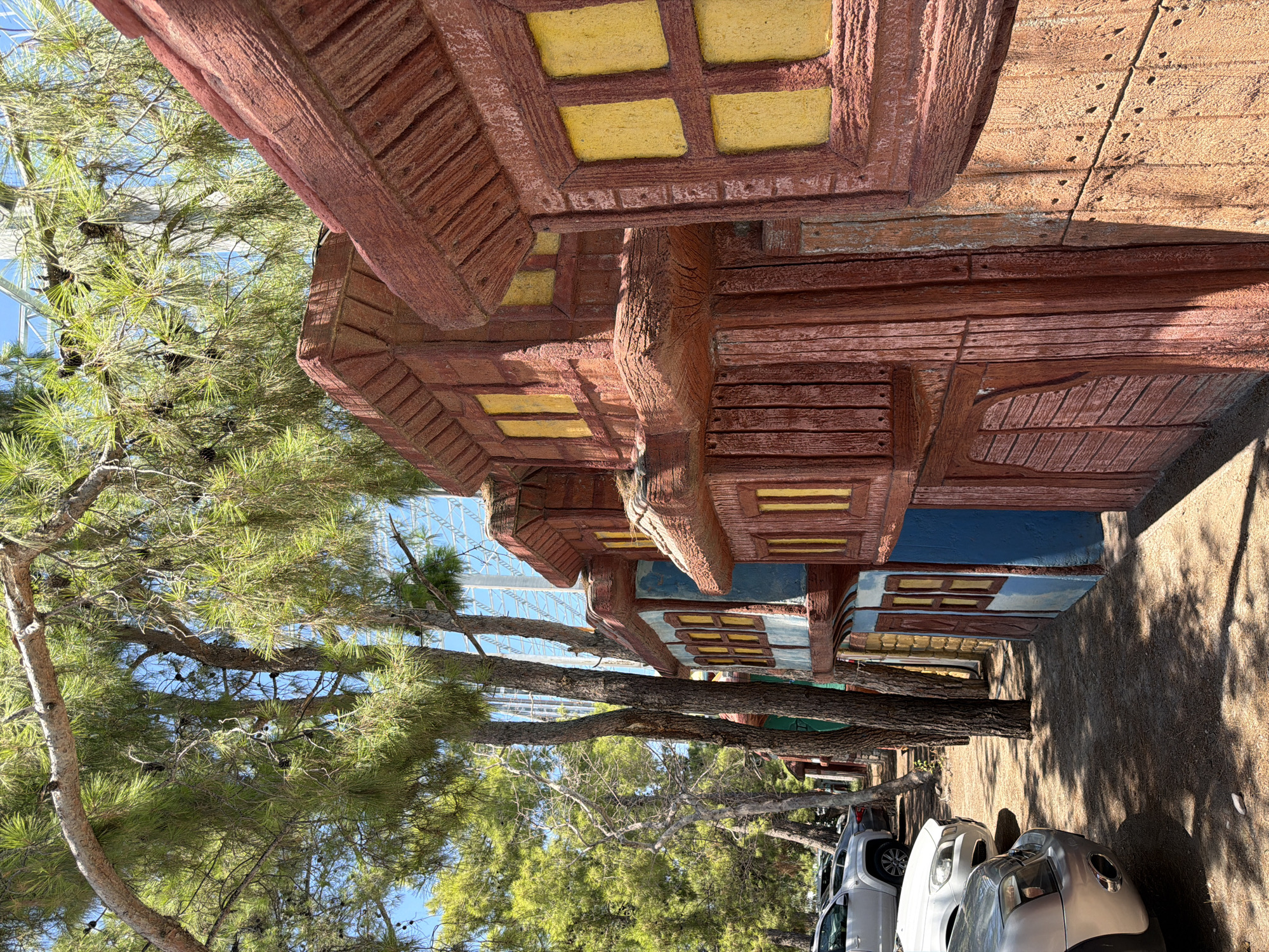 The image shows a row of artificial, miniature buildings resembling wooden houses, painted in red, brown, and blue. They have yellow window panes. The buildings are on a dirt path, with pine trees overhead casting dappled shadows. Several cars are parked on the left side of the path. In the background, a large, white, modern structure, possibly a building or a fairground attraction, is visible through the trees. The scene appears to be an outdoor, possibly recreational, area. The location is identified as Kemer, Turkey. There are no people visible in the image.