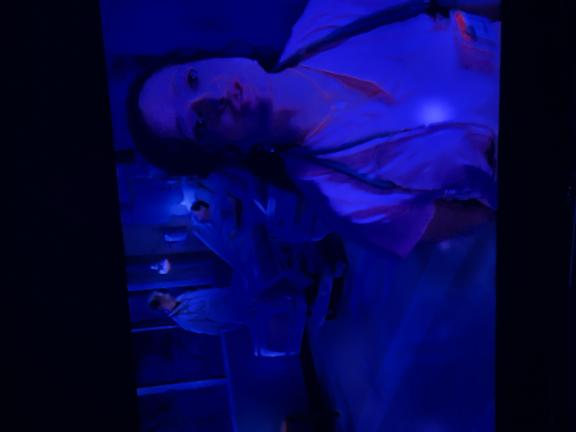 The image depicts a dimly lit hospital room bathed in a strong blue light, possibly from a blacklight. In the foreground, a person wearing a light-colored medical scrub top and a stethoscope is looking towards the camera. Their face is illuminated, revealing features and skin texture. In the background, another person in a white lab coat stands near a hospital bed. The bed is covered with a blue sheet, and a patient appears to be lying in it. Medical equipment, such as IV poles and monitors, might be present but are obscured by the lighting and distance. The overall atmosphere is sterile and somewhat eerie due to the dominant blue hue. There is no indication of the location being in Dobrich, Bulgaria, nor is there any information to suggest this.