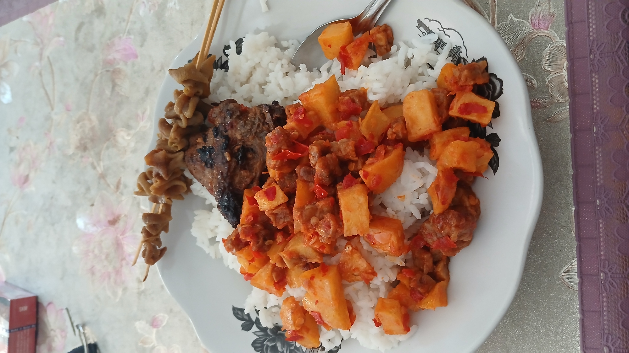 A white plate of food sits on a floral-patterned tablecloth. The plate contains white rice, a piece of grilled meat, and a dish of chopped potatoes and meat in a red chili sauce. Two skewers of cooked intestines are placed on top of the rice. A silver spoon rests on the right side of the plate. In the background, a portion of a box is visible. The scene appears to be in Sampang, Indonesia, based on the typical presentation of such meals.