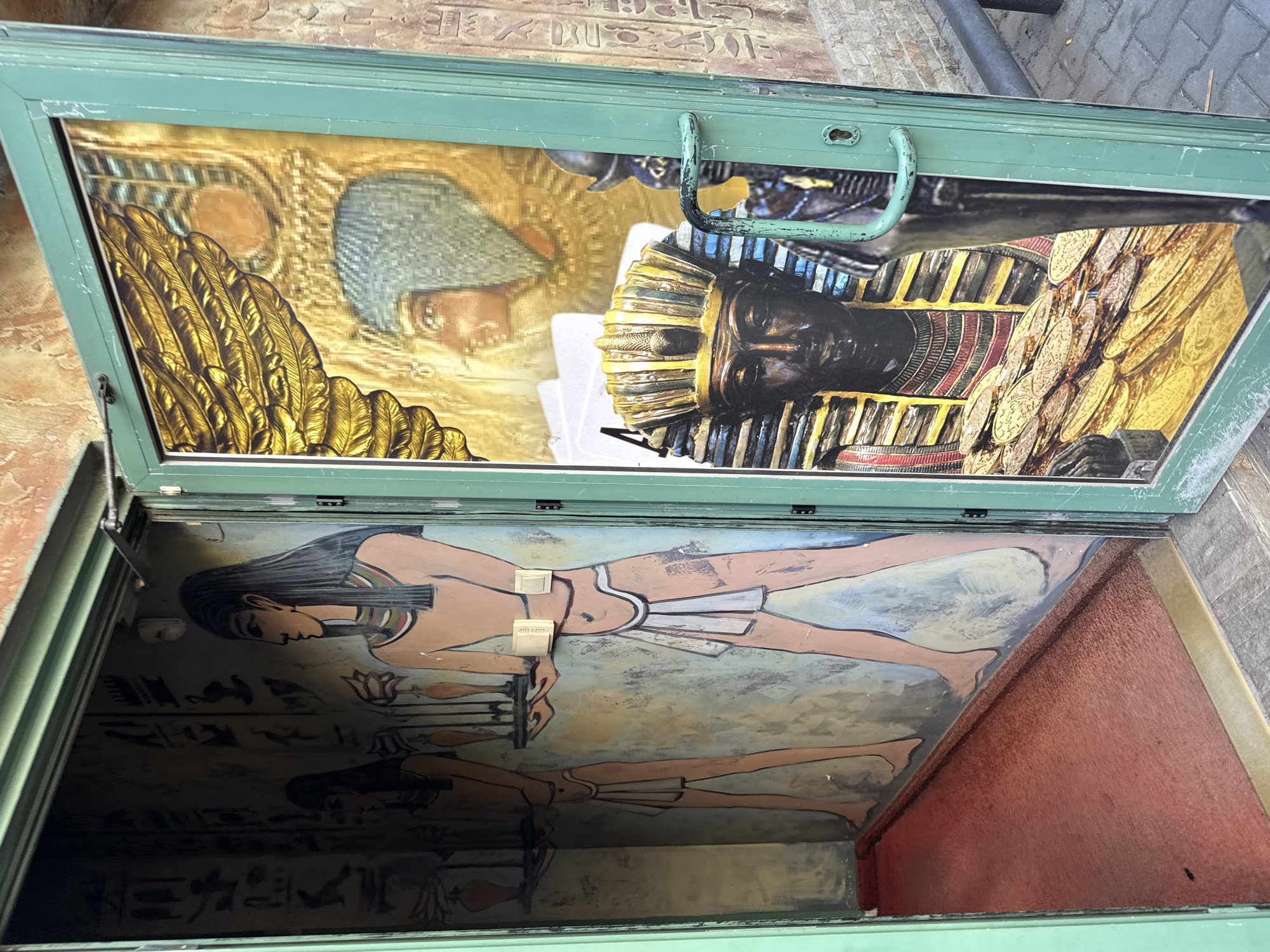The image shows an open doorway into an establishment decorated with Egyptian-themed murals. On the left, inside the doorway, a mural depicts two figures in profile, possibly ancient Egyptians, with hieroglyphics on the wall behind them. The floor inside is covered with a red carpet. The right side of the doorway features a glass pane displaying a mural of an ancient Egyptian sarcophagus, golden coins, and a pharaoh's headpiece. The exterior of the doorway is painted green, and the building's exterior walls are textured with a sandy color and stone accents. The location is specified as Stara Zagora, Bulgaria.