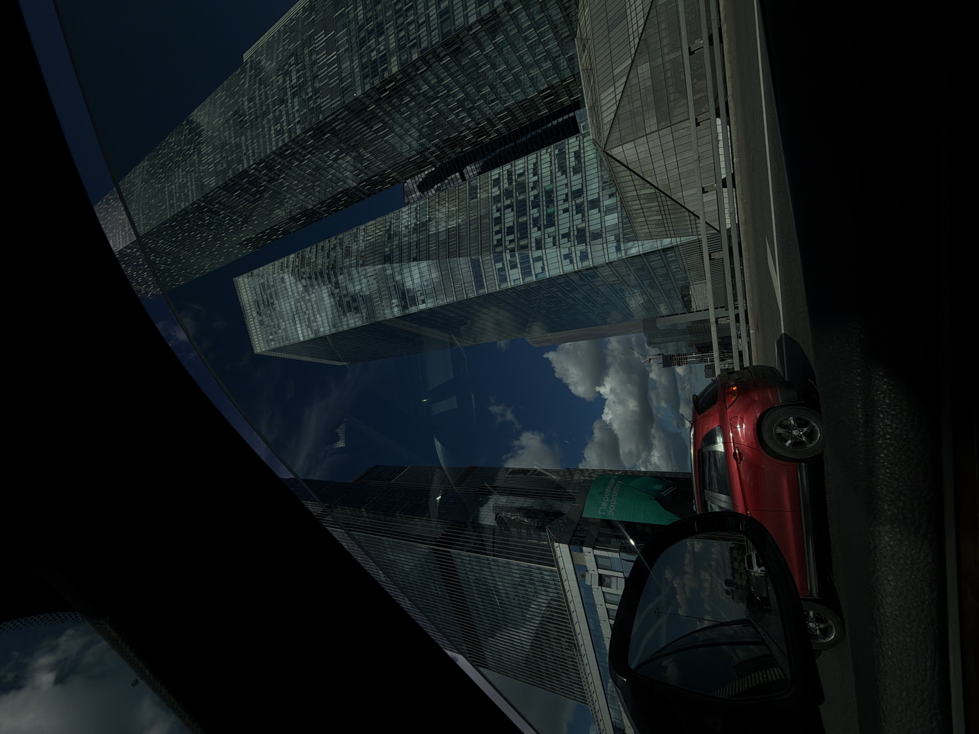 This media file depicts a view from inside a vehicle, looking out through the windshield and side mirror. The scene is dominated by a modern architectural landscape featuring several towering skyscrapers with reflective glass facades. The buildings are sleek and angular, reflecting the sky and clouds.
The perspective is such that the buildings appear to be upside down, suggesting a reflection in the car's window or a unique camera angle. The sky is a deep blue with scattered white clouds, indicating it is daytime, likely midday given the brightness and shadows. The weather appears clear and sunny.
In the foreground, a red car is visible, parked or moving alongside the road. It's a compact SUV-style vehicle. A side mirror of the car is also prominently featured, reflecting more of the cityscape and the sky.
One building facade has a large green advertisement with text in Cyrillic script. The visible text includes "Перевозка" and "золотая".
The overall impression is one of urban modernity and a dynamic cityscape, possibly captured during a drive through a business district or financial center in Moscow, Russia. The reflections add an element of visual complexity and depth to the scene.
