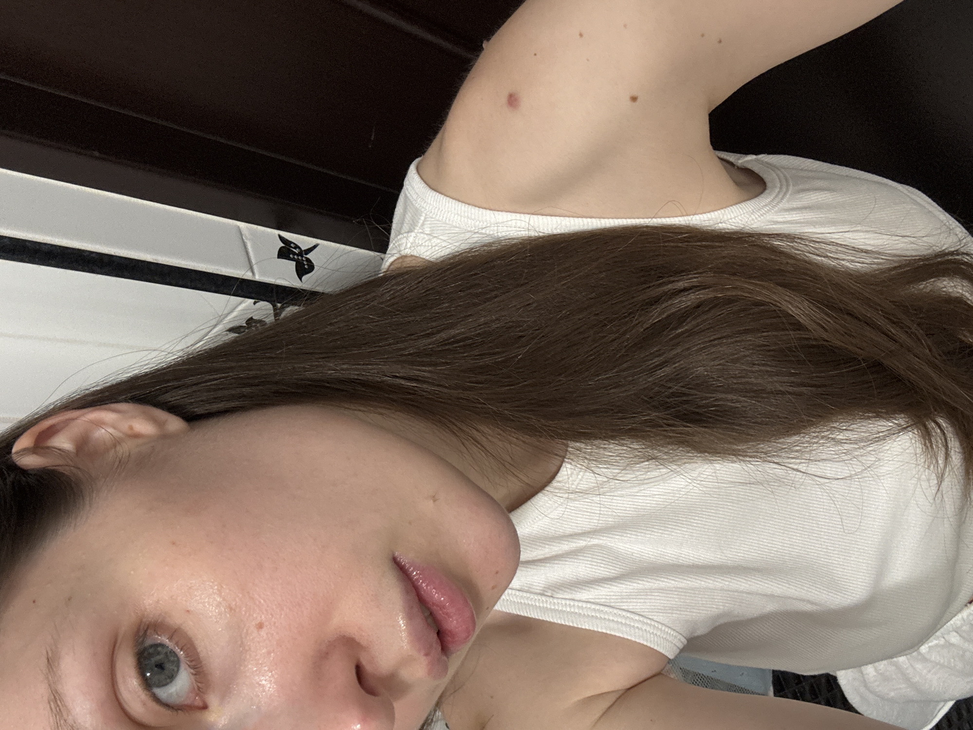 A close-up, high-angle shot depicts a person, likely a young woman, in an indoor setting. Her face, partially visible from the chin to just above the eyes, is angled towards the viewer, with one light blue-grey eye clearly visible. Her skin appears natural with some subtle moles and a slight sheen, possibly from natural oils or light moisturizer, and her lips are glossy pink. Long, dark brown hair cascades across her shoulder and chest, covering much of her white ribbed top.
On her visible shoulder, there is a small reddish mark along with a few moles. The background consists of a dark, possibly wooden or tiled surface above a white tiled or paneled wall. A distinctive black decorative element, resembling a stylized butterfly or bird adorned with small white dots (perhaps rhinestones), is visible on the white wall to the person's left.
The person appears to be posing for a selfie, given the intimate angle and focus on her face and upper body. The lighting is soft and even, suggesting an indoor environment without direct harsh light. No specific activity beyond posing for the image is evident, and her expression is neutral. There is no visible text. The setting is in Moscow, Russia.