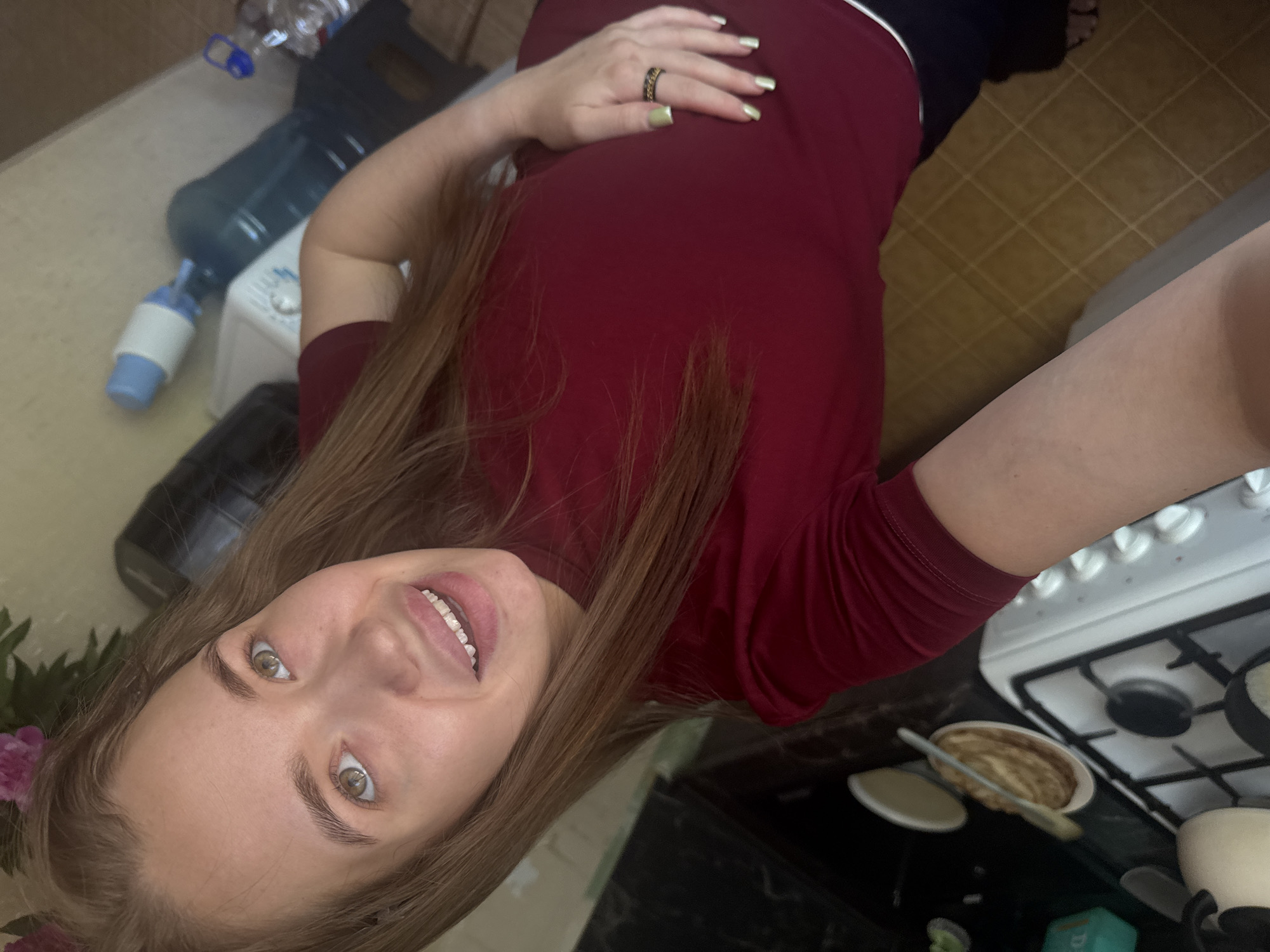 The media file shows a young woman lying on her side, holding her stomach. She is wearing a maroon t-shirt and has long, light brown hair. Her nails are painted a pale green, and she is wearing a ring on her right hand. The woman is smiling and looking at the camera. Her eyes are green and her eyebrows are well-defined.
In the background, there are several objects, including a large blue water cooler jug, a smaller white appliance, and a black object that could be a chair or couch. There is also a kitchen counter with a stove and some food items. The floor appears to be tiled.
The image is taken from a high angle, looking down at the woman. The lighting suggests it is daytime, possibly indoors in a kitchen. The woman's expression and relaxed pose suggest a casual and comfortable atmosphere. The presence of food on the stove could indicate that she is either preparing a meal or has just finished. The overall mood is relaxed and intimate. There is no visible text in the image. The location context is Chaikovsky, Russia.