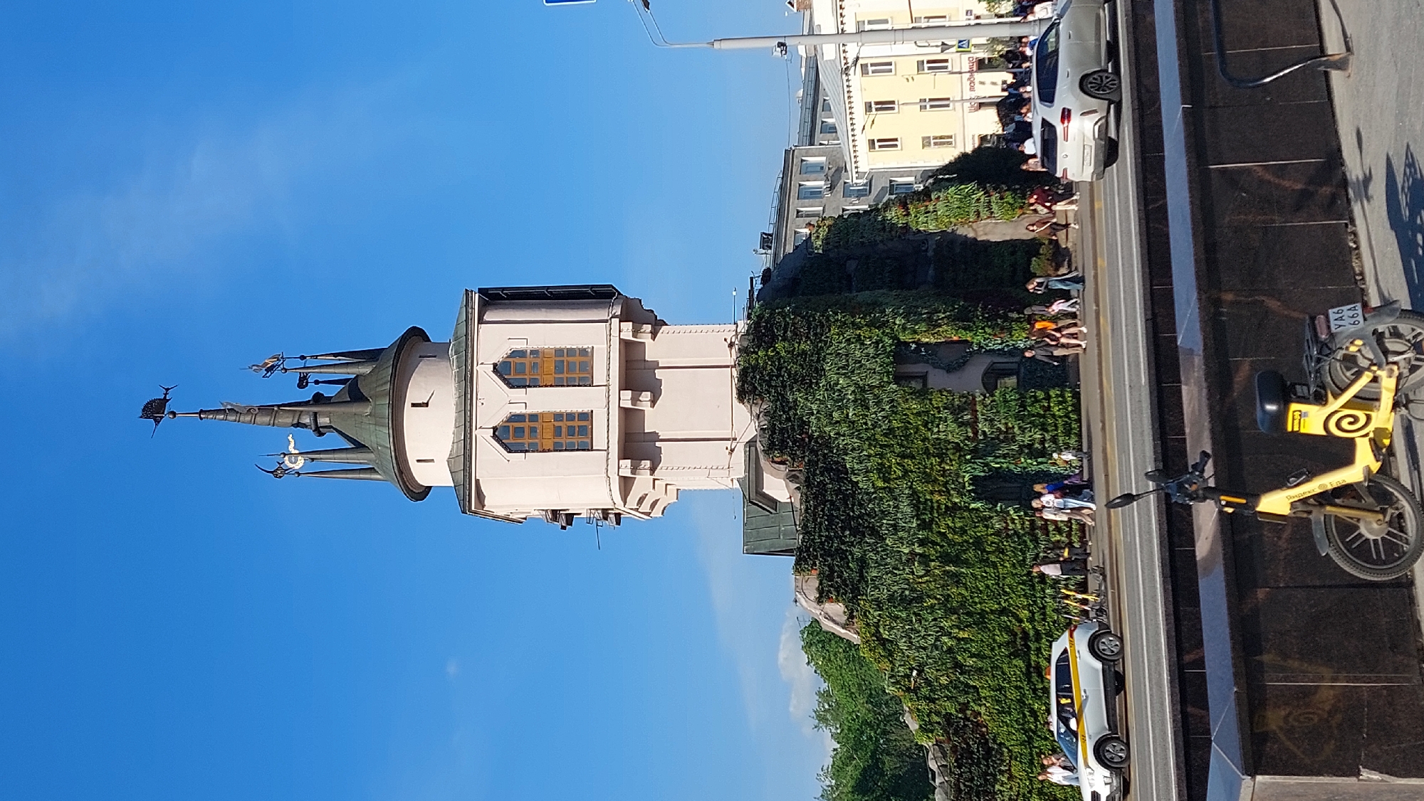 The image shows a low-angle view of the Central Children's Store at Lubyanka (formerly Detsky Mir) in Moscow, Russia, distinguished by its unique architecture with a tall, pointed tower.
The tower is light-colored, with windows set within its structure. The adjacent building is covered in green vegetation, and people are walking along the sidewalk next to a road with traffic.
On the lower right, there's a parked yellow scooter. Other vehicles, including a white car, are visible. The sky is bright blue and clear.