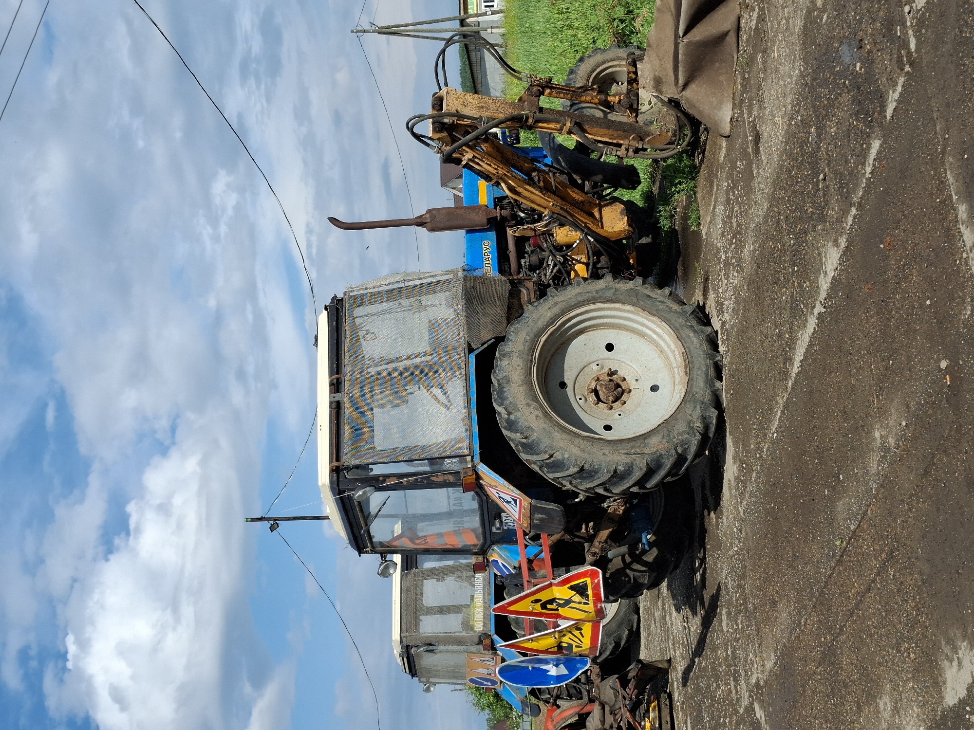 Two blue Belarus tractors are parked on a gravel surface. The tractor in the foreground has a yellow front loader attachment. Both tractors have enclosed cabs with metal mesh protection. Several road signs are attached to the side of the front tractor, including a blue directional arrow and two yellow triangular signs depicting construction workers. A tall exhaust pipe extends from the engine of the front tractor. The background shows a field of green grass, houses, and utility poles against a blue sky with white clouds. The image appears to be taken on a sunny day.