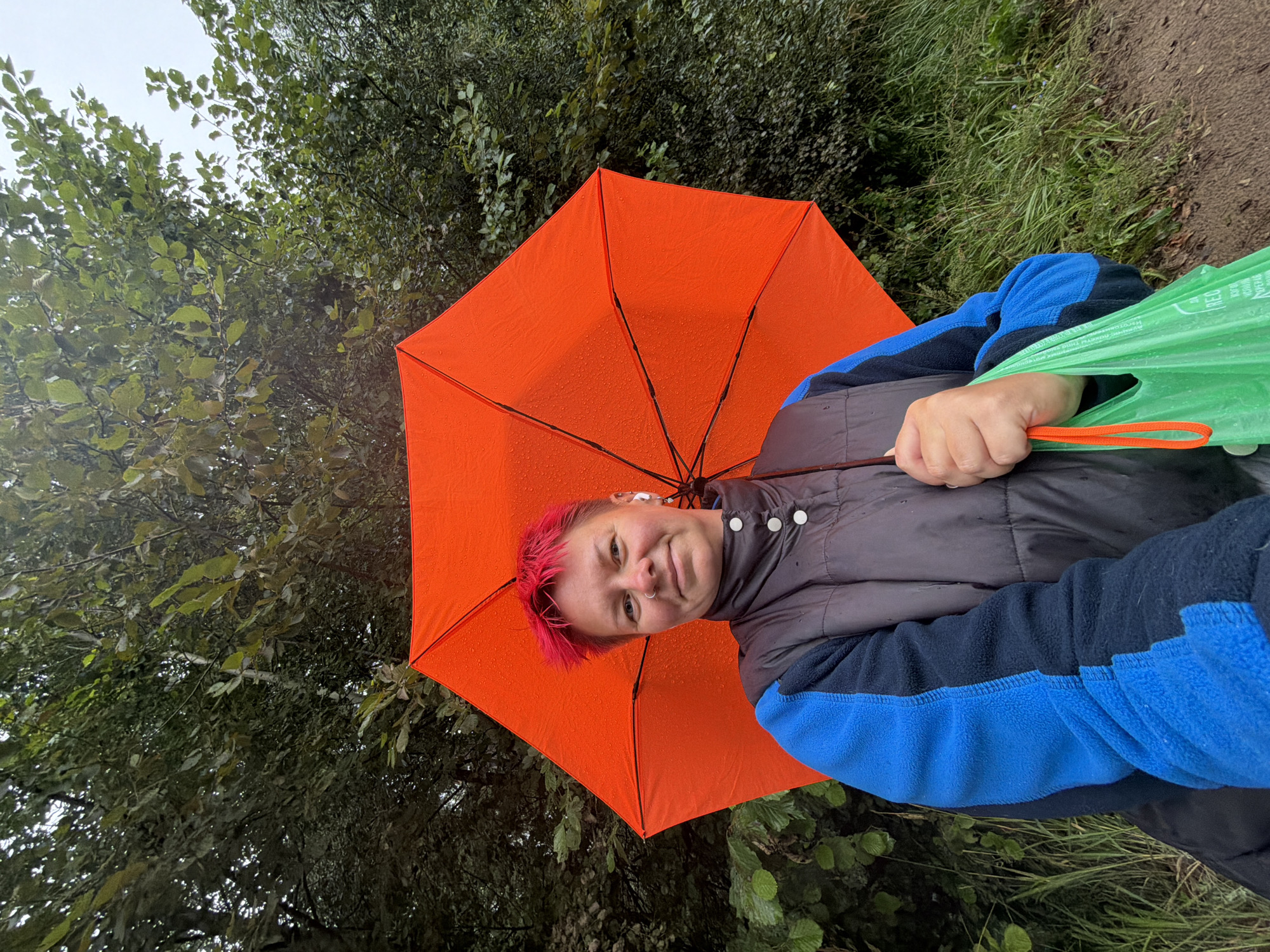 A person with short, bright pink hair is holding a large, open orange umbrella in the rain. They are wearing a dark puffer vest over a blue and black fleece jacket. They also have on white wireless earbuds, a nose ring, and a necklace with a pendant. In their right hand, they are holding a bright green plastic bag by its orange handles. The background consists of lush green trees and foliage, indicating an outdoor, natural setting, possibly a park or wooded area. The ground appears to be a dirt path. The scene suggests a casual outdoor activity in wet weather. There is no specific indication of the city of Chaikovsky, Russia in the image itself.