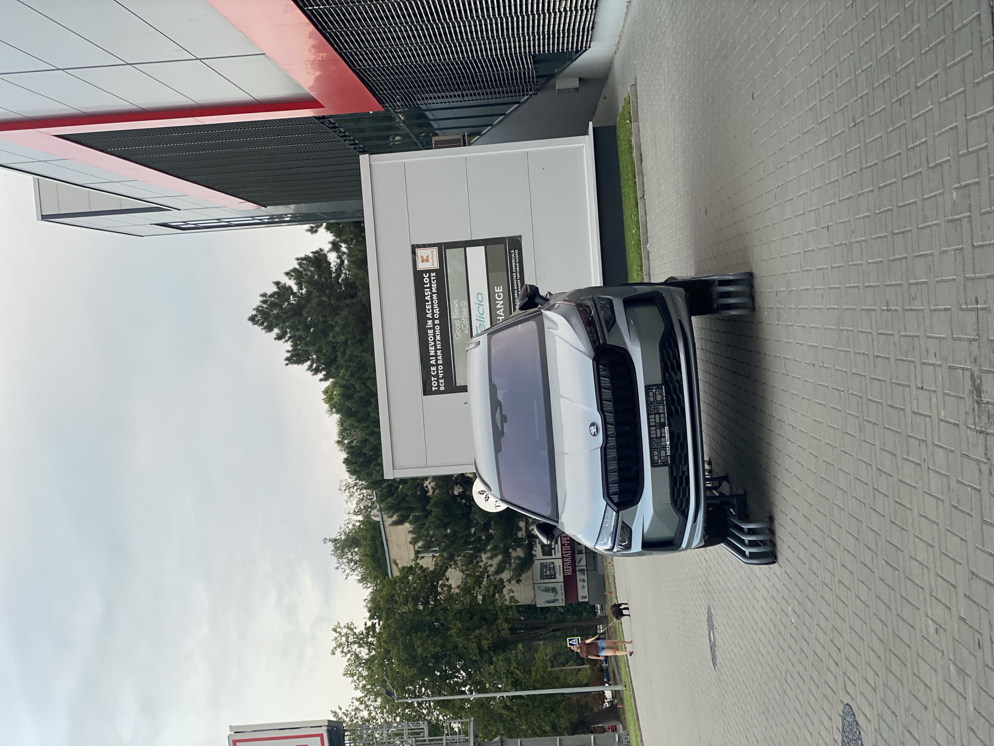 A silver Skoda Kodiaq SUV is parked on a paved lot outside a modern building. The car is positioned on a set of metal ramps that lift the front wheels. A woman is walking a small dog on a leash on the sidewalk in the background. The building has a large, segmented facade with red and grey panels and a dark grated section. A white kiosk with signage is situated next to the building. Trees and other buildings are visible in the distance under an overcast sky. The signage on the kiosk includes text in Romanian and Russian, with a "Kaufland" logo. The license plate of the car is visible and appears to be from Moldova.