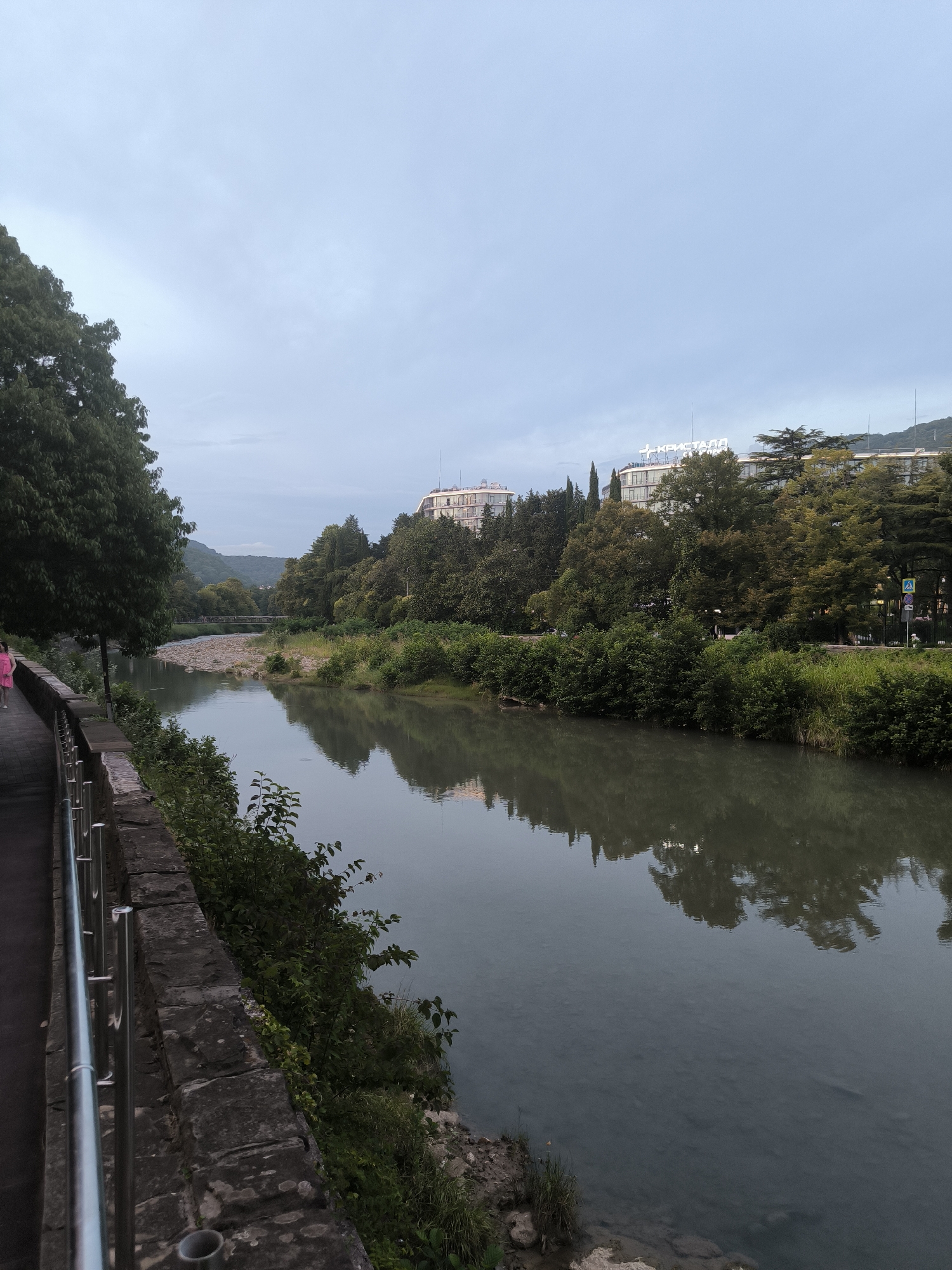 The image shows a wide, calm river flowing through a city. On the left bank, a paved walkway runs alongside the river, bordered by a stone wall and a metal railing. A young girl in a pink dress is walking on the walkway. Lush green trees and vegetation line both banks of the river. Across the river, several multi-story buildings are visible amongst the trees. One building prominently displays the word "КРИСТАЛЛ" (Kristall) in large white letters on its facade. In the distance, hills covered in trees rise against a cloudy, pale blue sky. The scene appears to be a riverside park or promenade in a city, likely Sochi, Russia, given the Cyrillic signage. The river reflects the surrounding trees and buildings, creating a serene and somewhat muted atmosphere.