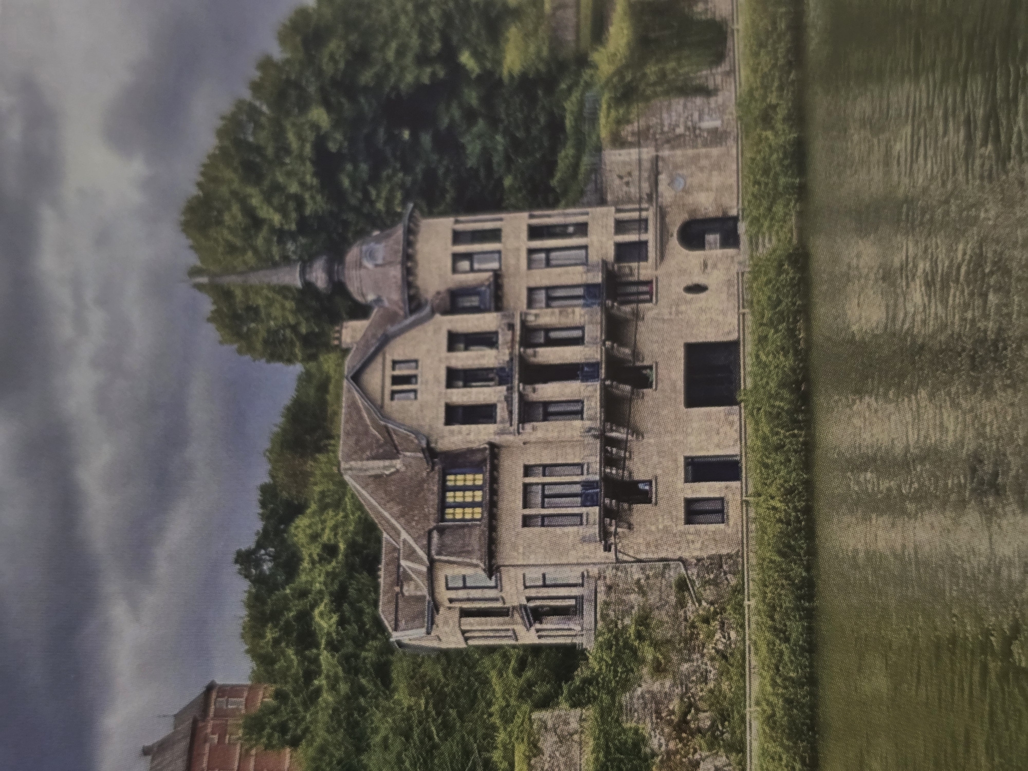 The image presents a grand, multi-story building, reminiscent of a chateau or large historical residence, set against a backdrop of lush natural scenery. The building, constructed from light-colored stone or brick, exhibits a weathered facade with numerous rectangular windows, some featuring dark frames, and several small balconies or ledges. A distinctive architectural element is a round turret topped with a pointed spire roof on the left side of the structure. The rooflines are complex, indicating multiple sections and gables.
Dense green trees flank and surround the building, creating a vibrant natural frame. To the right of the building, separated by a narrow strip of foliage, lies a large expanse that appears to be either a body of water with subtle ripples or a cultivated field with distinct textures.
The sky is overcast with dark, heavy clouds, suggesting a cloudy day, possibly with impending rain. The lighting is diffused and even, characteristic of such weather conditions. No people or discernible activities are present, rendering the scene tranquil and static. There is no visible text within the image. Based on the provided context, the location of this scene is Sofia, Bulgaria.