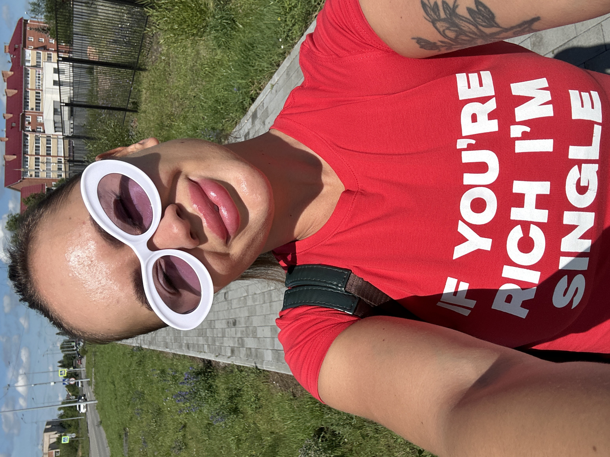 A woman is taking a selfie outdoors on a sunny day. She is wearing large white oval sunglasses and a red t-shirt with white text that reads "IF YOU'RE RICH I'M SINGLE". She has full, glossy lips and her hair is pulled back. She is carrying a dark green backpack. The background shows a paved sidewalk, a grassy area with some purple flowers, a black metal fence, a road with traffic, and buildings in the distance. Based on the architecture and signage (road signs in Russian, buildings with distinctive red brick roofs and window styles common in Russian apartment blocks), the scene is likely in Yuzhnouralsk, Russia. The woman's pose and expression suggest she is confident and perhaps playful. The overall atmosphere is casual and summery.