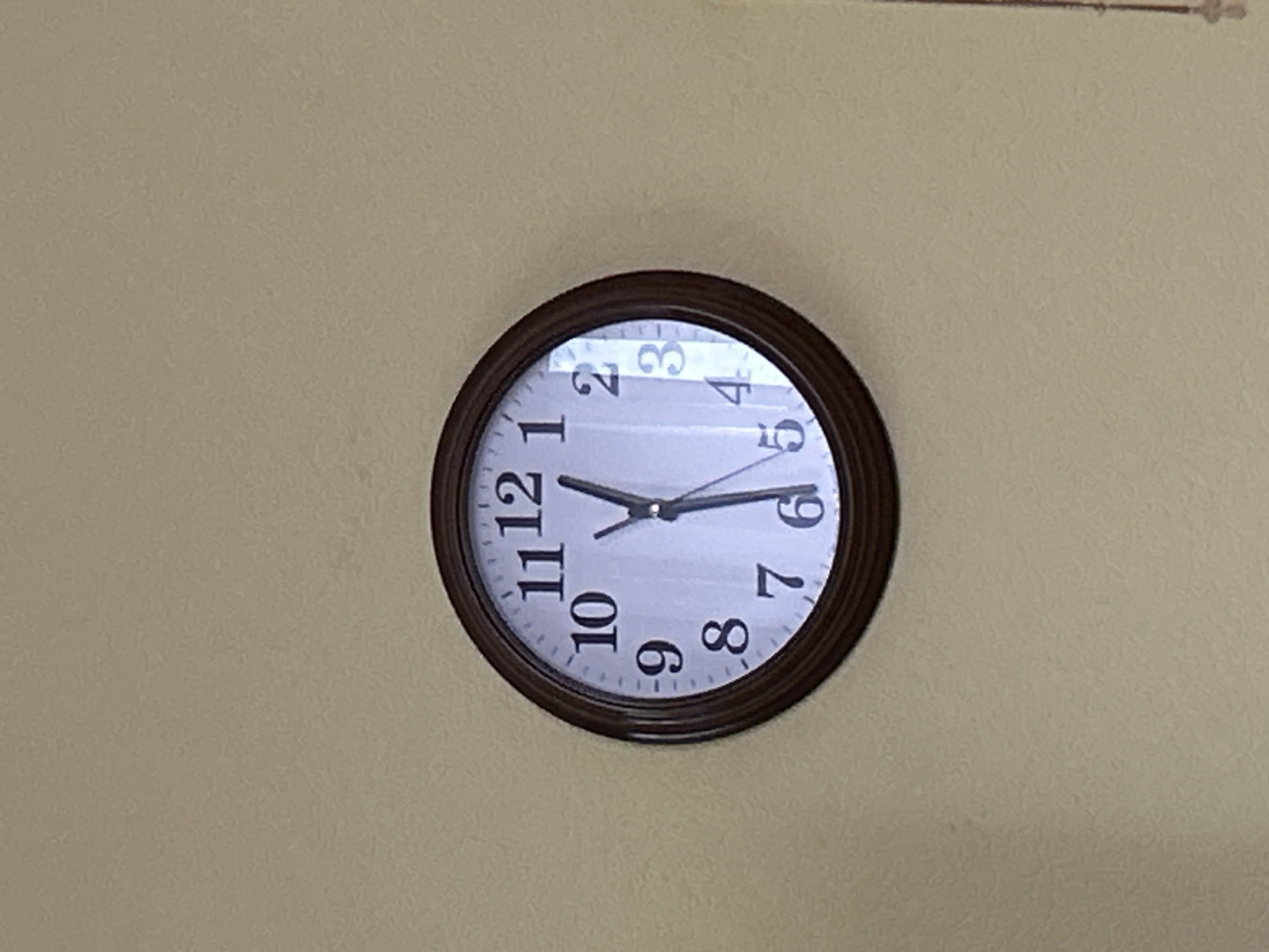 A brown-framed analog clock with a white face hangs on a beige wall. The clock shows the time as approximately 12:59. The numbers on the clock are black and clearly visible, ranging from 1 to 12. The hour hand is very close to the 12, and the minute hand is pointing at the 12. The second hand, a thin black line, is just before the 12. There is a reflection on the clock face, obscuring some details. The wall appears to be textured. The location is Oktyabrskoye, Russia.