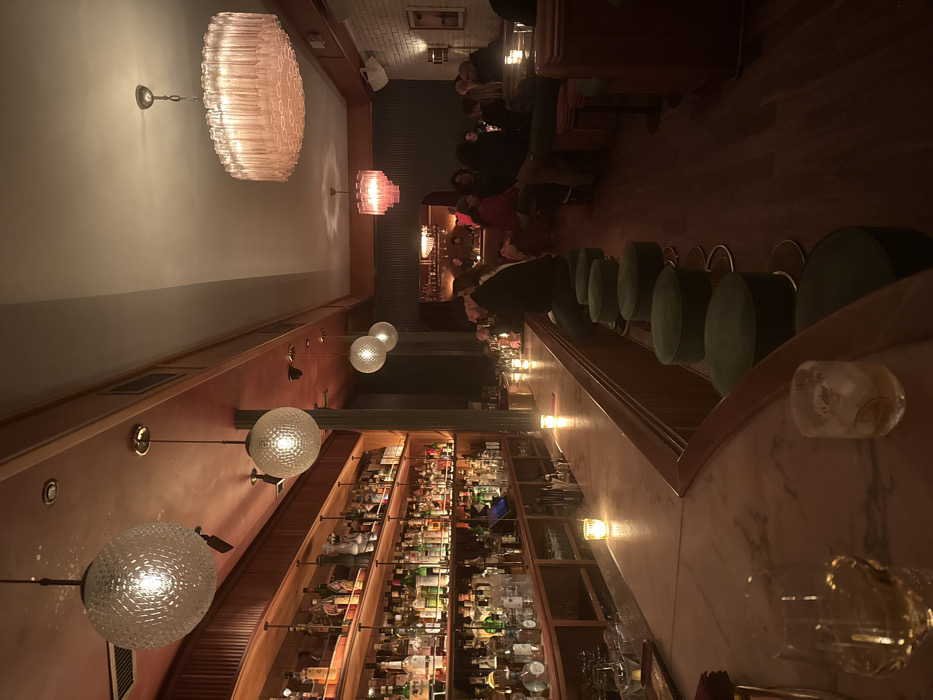 This image depicts the interior of a dimly lit bar in the city of New York, United States of America. The scene focuses on a long, curved marble-topped bar counter extending from the foreground to the midground on the left. Several small, lit candles in clear glass votives are spaced along the bar surface. In the immediate foreground, two glasses containing amber-colored beverages with ice are visible on the counter; one large glass is on the bottom left, and a smaller one is central.
A row of dark green, velvet-upholstered bar stools with golden bases is positioned along the bar. Several patrons are seated at the bar, one prominently visible from the back in a dark jacket. Behind the bar, a multi-tiered wooden shelving unit is densely stocked with various liquor bottles, illuminated by internal strip lighting.
The ceiling above the bar is reddish-brown, featuring multiple spherical, textured glass pendant lights with warm glow. Further to the right, a section of the ceiling is a lighter color, from which a large, ornate chandelier with numerous pink-tinted glass tubes hangs. A smaller, tiered pink chandelier is visible deeper within the space. The walls include dark, possibly fluted paneling and a section of white brick.
In the background, a seating area with dark booths or benches accommodates additional patrons, their faces largely obscured by the low light. An arched opening reveals another dimly lit area deeper inside the establishment. The overall environment suggests an upscale, intimate evening setting.