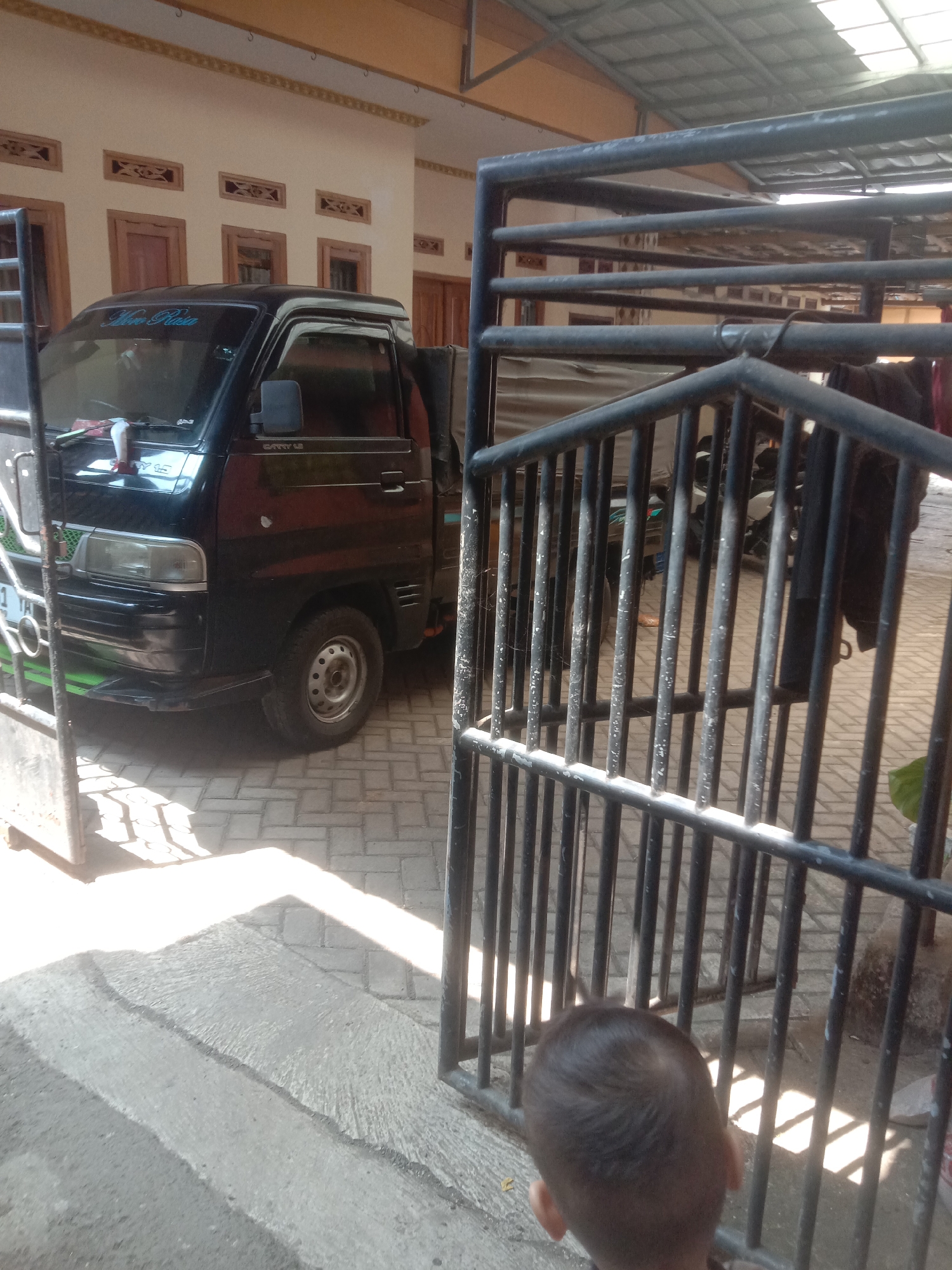 The image depicts a black Suzuki Carry pickup truck parked on a paved surface in what appears to be a residential or commercial area in Bojong, Indonesia. The truck has a green accent on its front bumper and a tarp covering its bed. A metal gate with vertical bars is in the foreground, partially obscuring the view of the truck and the environment behind it. To the right of the gate, a young child with dark hair is visible from behind, looking into the scene. The background shows a building with a light-colored facade and decorative window elements. Sunlight is casting strong shadows on the ground. The overall scene suggests a typical outdoor setting in Indonesia, possibly a driveway or an open yard.