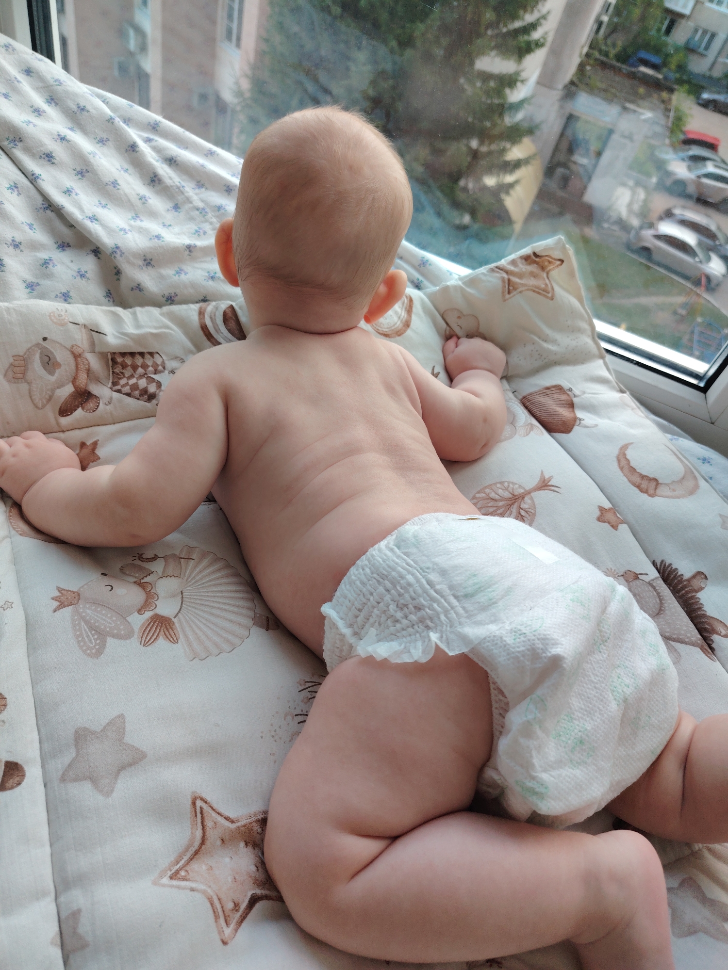 The image shows a baby lying on its stomach on a patterned blanket near a window. The baby is wearing a diaper and is facing away from the camera, looking out the window at a residential area with buildings and parked cars. The scene is set indoors, likely in a home, with the view outside suggesting an urban or suburban environment. The blanket has a pattern of stars and cartoon animals. The lighting indicates it is daytime. There are no other individuals visible in the image.