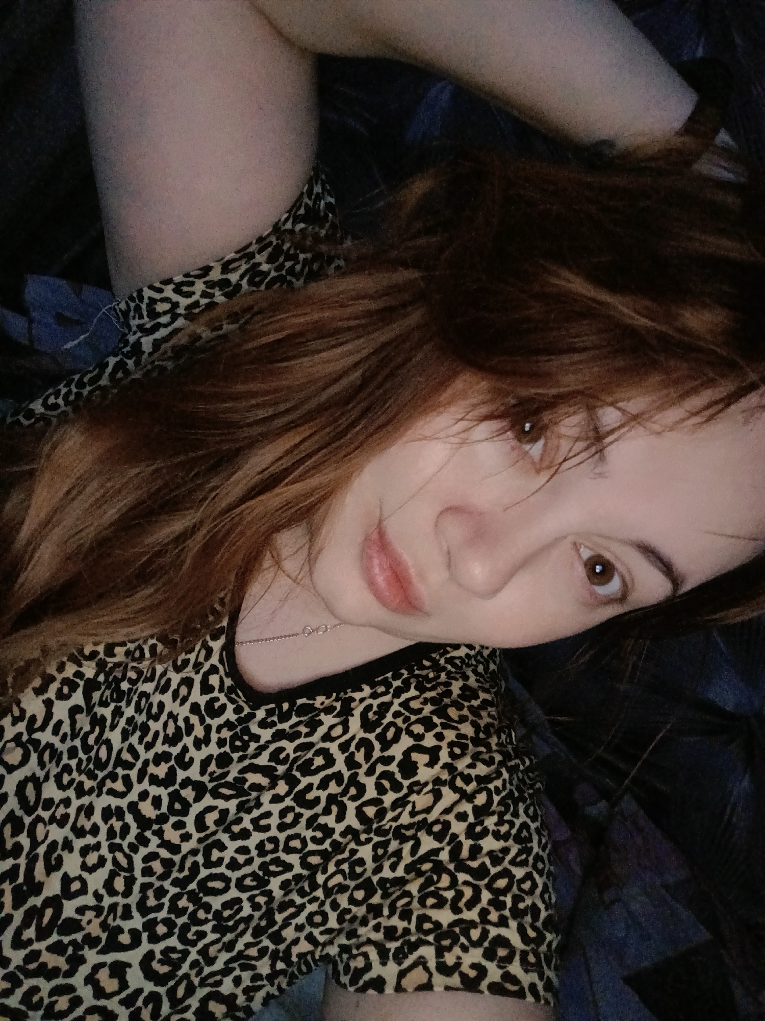 A woman with light skin and reddish-brown hair is lying down. She is wearing a leopard-print shirt and a thin silver necklace. She is looking directly at the camera. Her right arm is raised above her head. The background is dark, but there is some blue fabric visible. There is no evidence or indication that this image was taken in Chaikovsky, Russia.