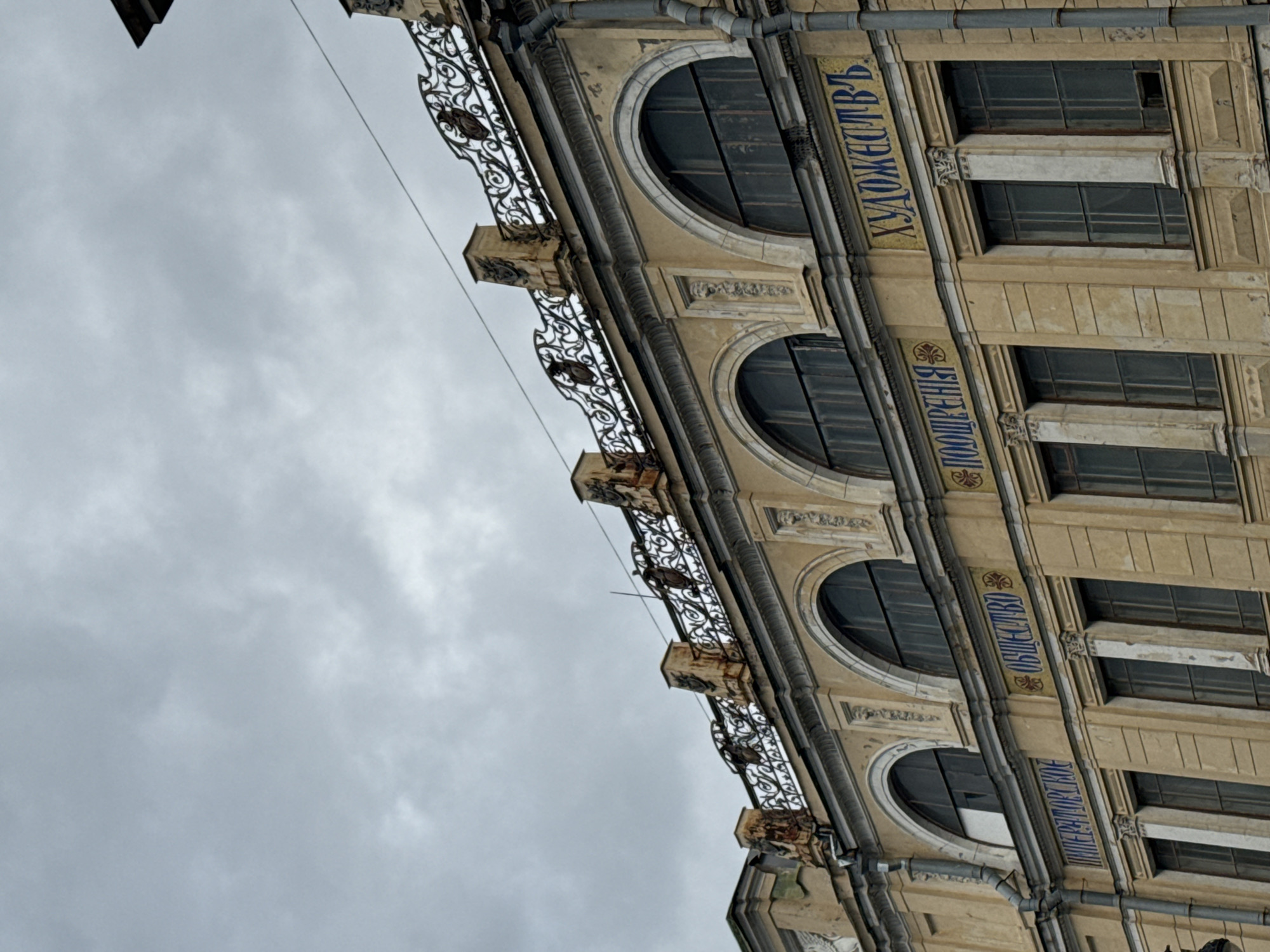 The image shows the facade of a historical building in Saint Petersburg, Russia, under a cloudy sky. The building is constructed in a classical architectural style with ornate details. The facade is painted a light yellow, and features arched windows on the upper level, with decorative ironwork above them. Along the facade, there are inscriptions in blue Cyrillic lettering. The inscription on the left reads "ИМПЕРАТОРСКОЕ", the middle reads "ОБЩЕСТВО ПООЩРЕНИЯ", and the right reads "ХУДОЖЕСТВЪ". These inscriptions translate to "IMPERIAL SOCIETY FOR THE ENCOURAGEMENT OF ARTS". The building appears to be well-maintained but shows signs of age, with some peeling paint and weathering. The overall atmosphere is somber, likely due to the overcast weather. No people are visible in the image.
