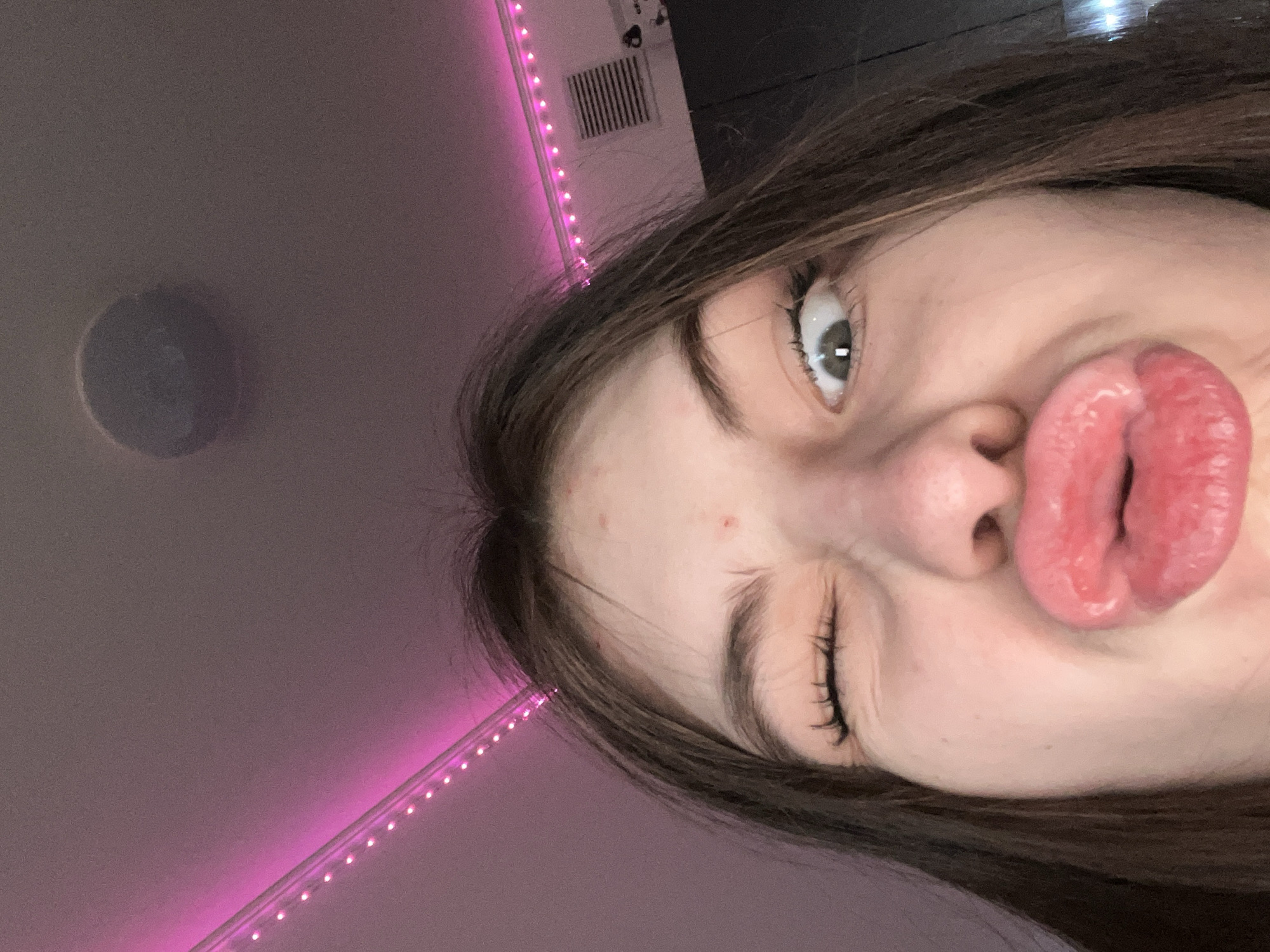 A young woman is close-up, making a kissing face with one eye squinted shut. Her skin appears fair, with a few small blemishes. She has dark hair and light-colored eyes. The background includes a light pink ceiling with a line of LED lights, an air vent, and a round ceiling fixture. The location is not discernible from the image.
