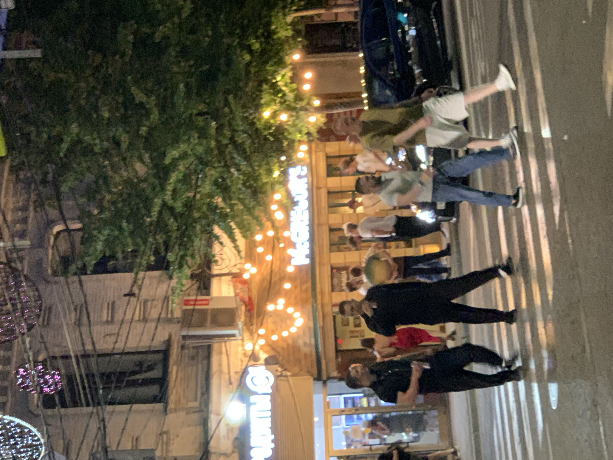 In a city at night, a group of people are crossing a street at a pedestrian crossing. The street is illuminated by decorative string lights hung from buildings and streetlights. One building has a sign with Cyrillic lettering and another sign that reads "McGregor's". A dark blue car is parked behind the pedestrians. The weather appears to be clear, with some reflections on the wet pavement of the street.