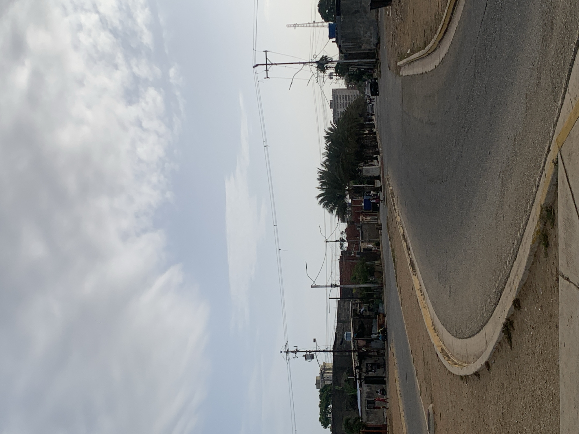 An outdoor, daytime shot depicts a street scene under a bright, overcast sky. The image is oriented vertically but appears to be a landscape captured on its side, showing a road curving from the bottom right towards the middle left of the frame.
The asphalt road surface, appearing somewhat worn with visible cracks, features a clear yellow painted line on its concrete curb. This curb separates the road from a dusty, unpaved strip of ground. Further to the left, another road or path is partially visible.
Along the main street, numerous utility poles are strung with a dense network of power lines, crossing the sky. A prominent line of lush palm trees runs parallel to the road, partially obscuring a mix of buildings behind them. These structures range from what appear to be residential houses to larger, multi-story buildings in the distance, some showing signs of disrepair or ongoing construction. A dark-colored vehicle is visible on the street further down.
The sky occupies a significant portion of the upper left, filled with soft, diffuse light-grey and white clouds, suggesting an overcast or partly cloudy day. No specific human activity is clearly discernible, nor is any legible text visible within the frame. The overall impression is of an urban or semi-urban environment in a warm climate.