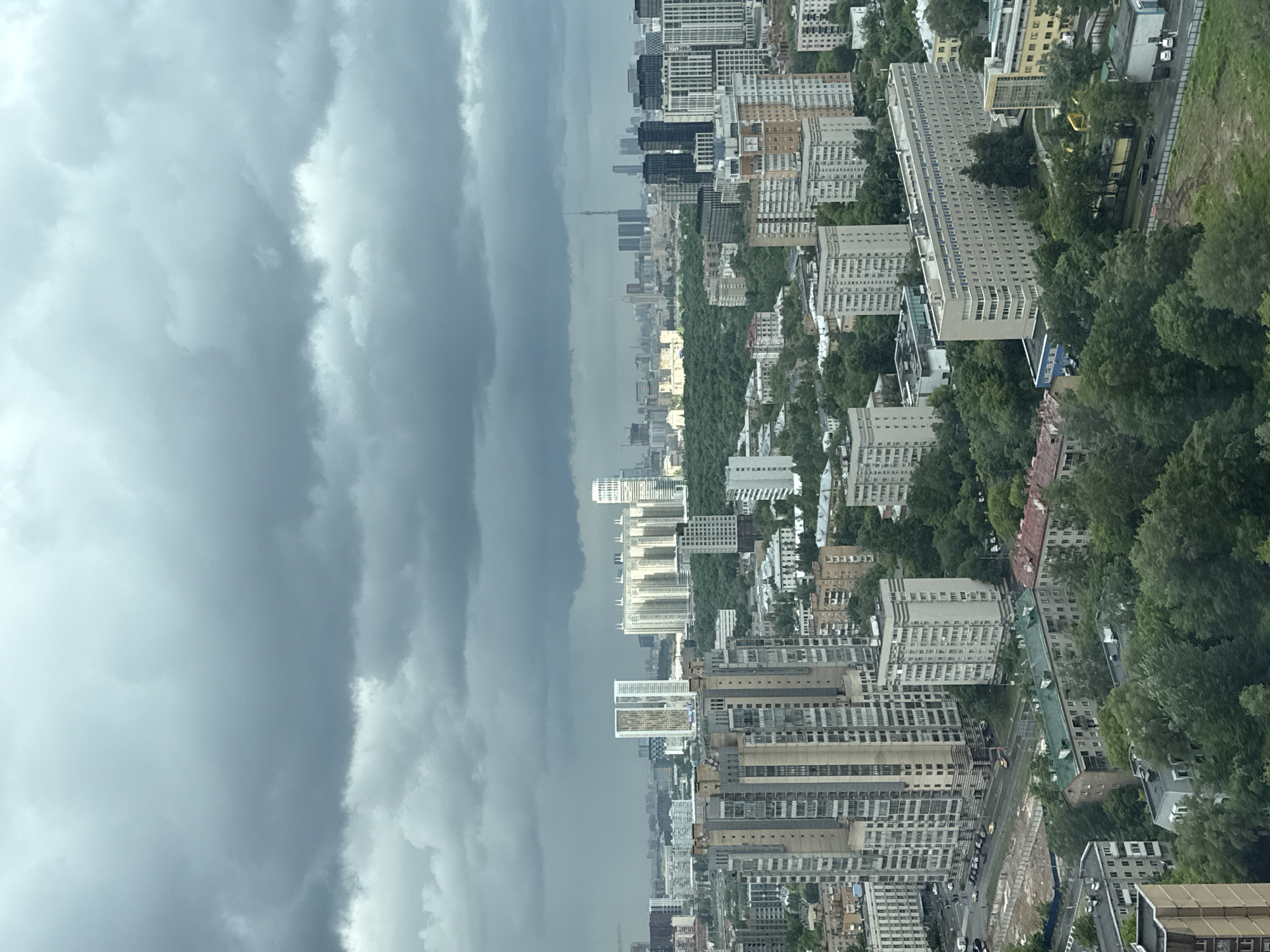 The image captures an aerial view of Khimki, Russia, under a sky dominated by dense, ominous storm clouds. The urban landscape consists of a mix of high-rise apartment buildings and office blocks interspersed with green spaces, notably parks and tree-lined avenues. The architecture is predominantly utilitarian, featuring repetitive designs and a mix of gray, beige, and white facades. In the distance, a tall communication tower or radio tower is visible on the horizon. The environment suggests an impending storm, with the dark sky casting a subdued light over the city. Traffic appears minimal on the roads below, and there are no visible pedestrians or signs of human activity.