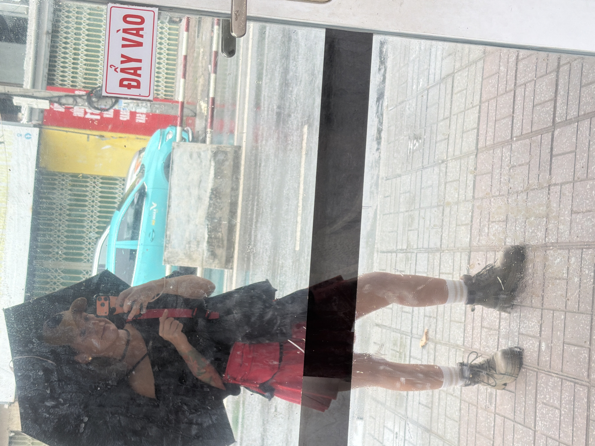 A person holding a black umbrella takes a selfie in a reflection on a shop window. The person is wearing a black off-the-shoulder top, a red pleated skirt with a black underskirt, white socks, and dark boots. They have a red fanny pack around their waist and a choker necklace. In the background, through the window, a turquoise car is visible on a wet street, suggesting it is raining. A yellow building and a red sign with Vietnamese text "ĐẨY VÀO" (Push In) are also visible. The reflection shows the person standing on a tiled sidewalk. The scene is likely on a street in Nha Trang, Vietnam, given the Vietnamese text on the sign.