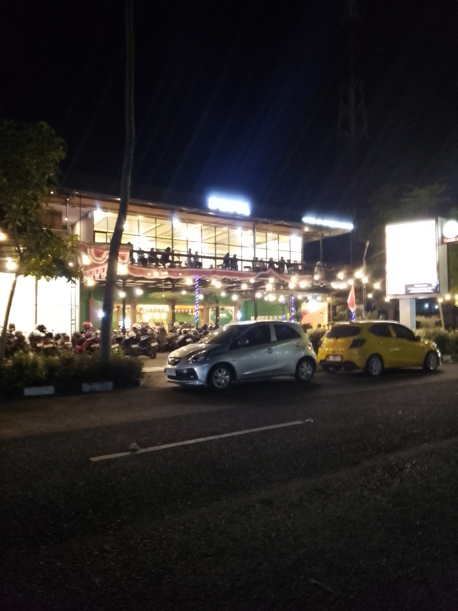 A night scene depicts a multi-level building with an open-air upper floor balcony where several people are seated, suggesting a restaurant or cafe. The building is illuminated by numerous string lights and interior lights. In front of the building, a row of motorcycles is parked. Two cars are visible on the paved road in the foreground; a silver hatchback is on the left, and a yellow hatchback is to its right. The road appears to be asphalt with painted white lines. The background is dark, with the faint outline of a tower or mast visible against the night sky. Trees and foliage are present to the left of the building. This scene is likely located in Lamongan, Indonesia, given the architecture and the presence of motorcycles.