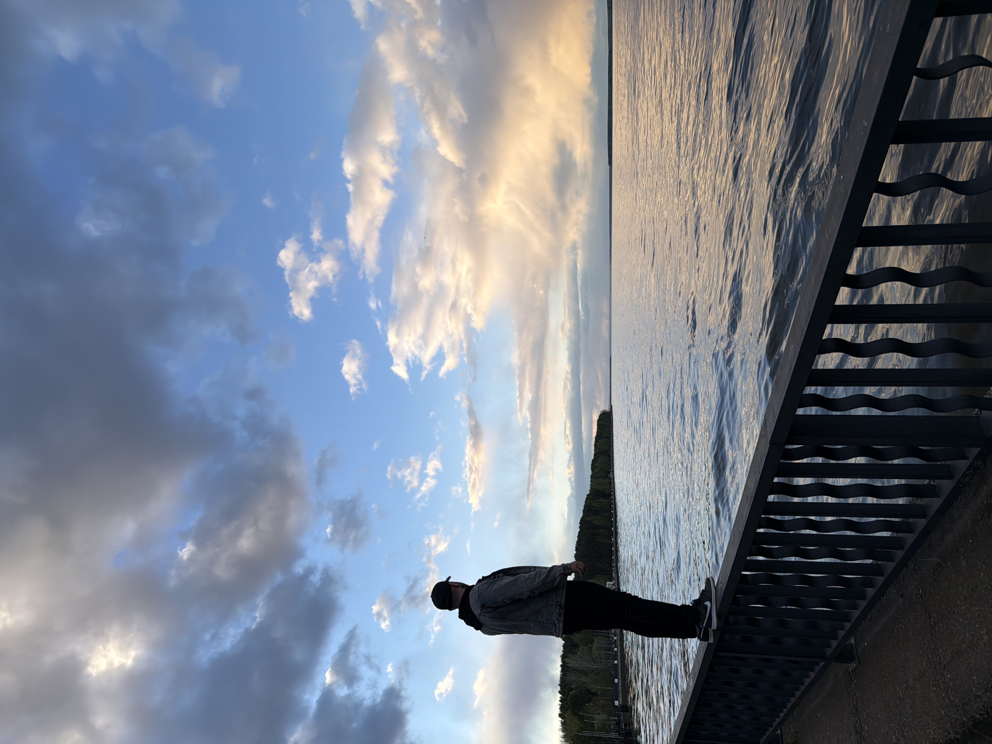 A man stands on a bridge in Chaikovsky, Russia, facing a body of water. He is wearing a black baseball cap, a gray denim jacket, black pants, and white sneakers. The water is rippled and reflects the sunlight and clouds. The sky is blue with a mix of dark and light clouds. A forested area is visible in the distance across the water. The bridge has a dark gray metal railing with a wavy design.