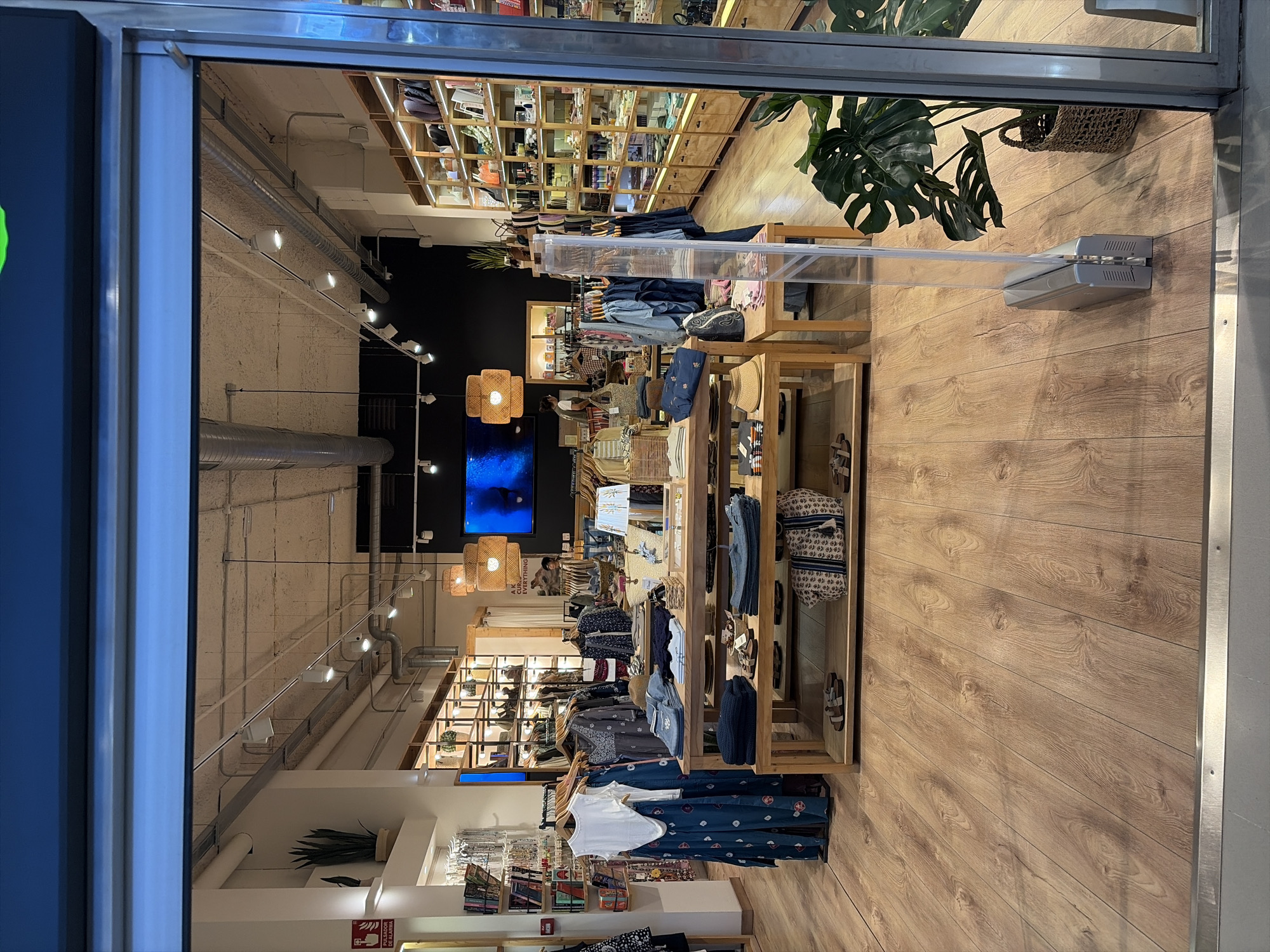 The image shows the interior of a retail store, likely a clothing boutique, located in Antigua, Spain. The store has a modern, minimalist design with natural wood finishes and warm lighting. In the foreground, there are wooden display tables showcasing folded clothing items such as sweaters, jeans, and shirts, as well as straw hats and tote bags. Various garments are also displayed on hangers on racks to the left. In the background, shelves are stocked with bags, books, and other merchandise. A large screen is mounted on the back wall displaying an underwater scene. The floor is made of light-colored wood planks. Natural light streams in from an open entrance. A large potted plant with broad leaves is visible to the right of the entrance.