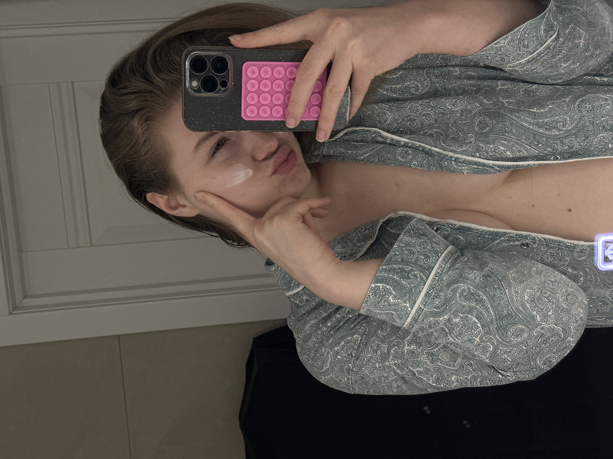 A female subject is shown taking a mirror selfie indoors. She has light brown hair and is wearing an unbuttoned, light blue or teal pajama-style shirt adorned with a white paisley pattern and white piping, revealing her upper chest and torso. A white cream or product is visible on her left cheek, which she touches with her left index finger while exhibiting a pouting facial expression. She holds a dark-colored smartphone, identified by its triple camera array and glittery dark case, in her right hand. A pink rectangular pop-it fidget accessory with multiple circular bubbles is affixed to the back of the phone case. The background, visible in the mirror's reflection, includes a plain light-colored wall to the left and a white paneled door to the right. A dark, indistinct object is partially visible on the far left of the reflection. The specific location within Kazan, Russia, cannot be determined from the visual information provided in the image, but the setting appears to be a private interior space.