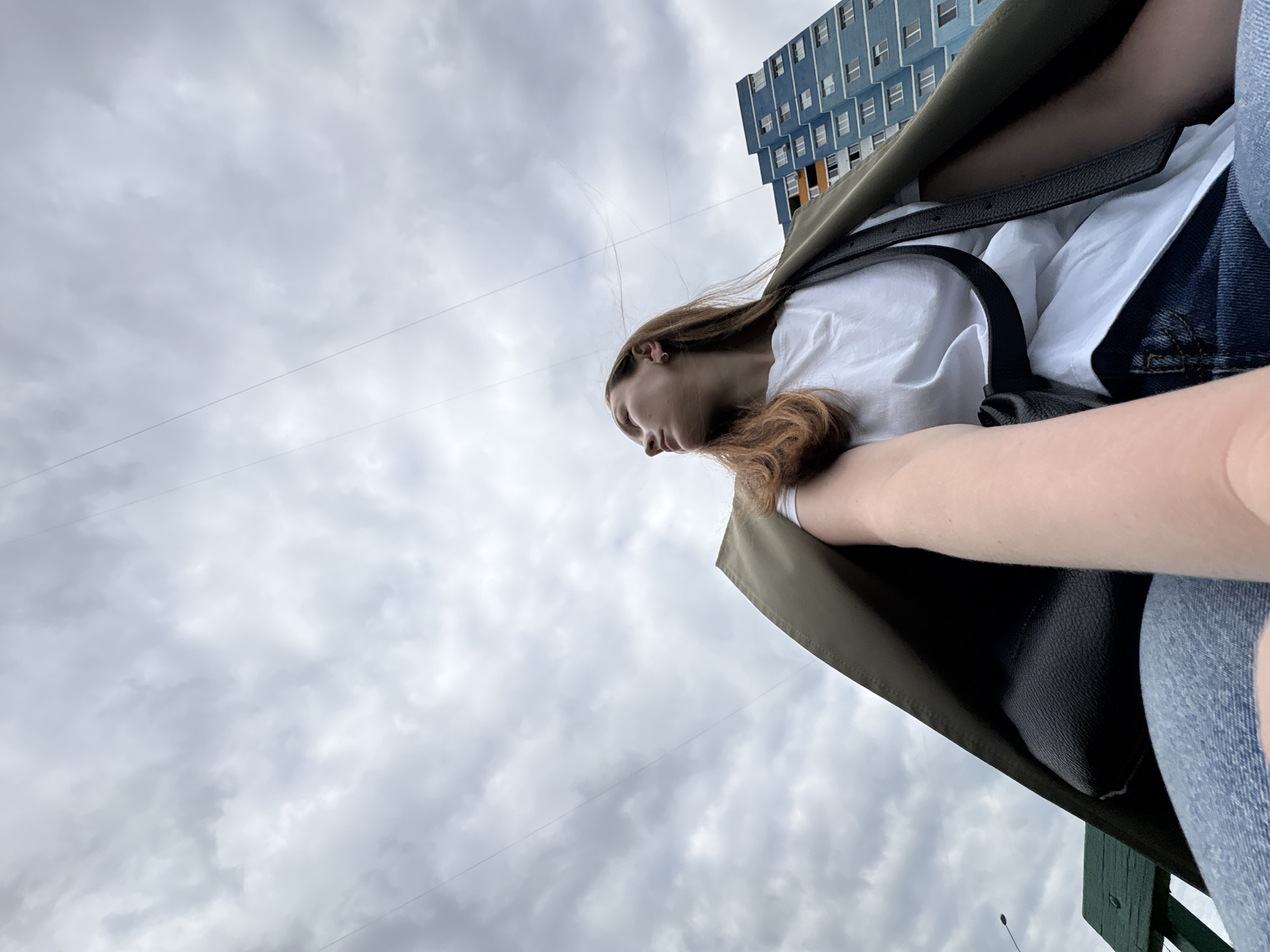 A low-angle, medium shot captures a young woman from the chest up, seated on a green bench, looking away from the camera toward the left. She has long, auburn hair and is wearing a white t-shirt, blue jeans, and an olive-green jacket draped over her shoulders. A black purse with a strap is visible. Behind her, a tall, blue apartment building with many windows dominates the background under a cloudy, overcast sky. Thin wires are visible against the sky, suggesting a utility line or antenna. The location is implied to be Izhevsk, Russia, based on context not present in the image itself.
