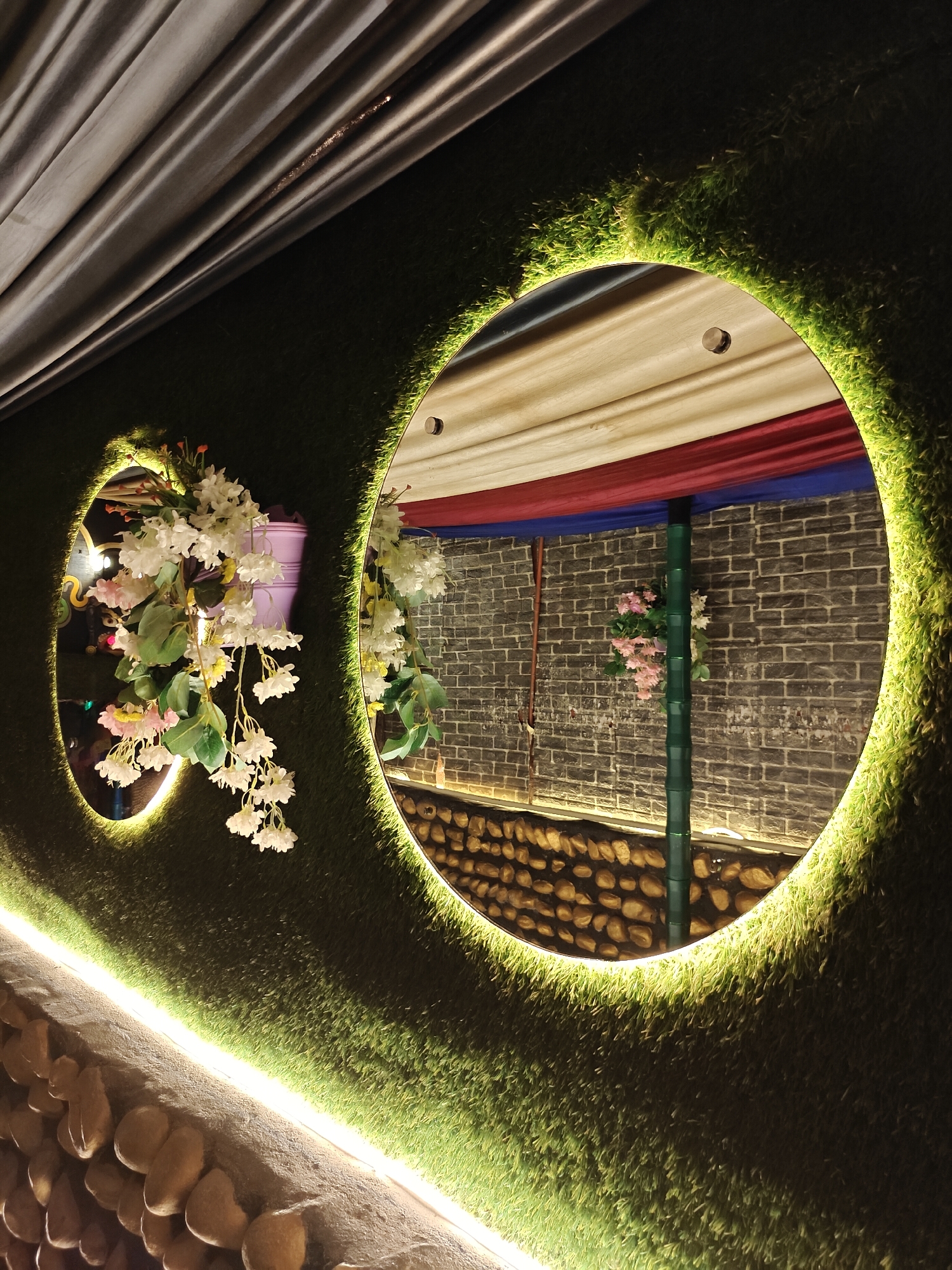 This image shows a decorative wall featuring two oval mirrors embedded in artificial grass. The mirrors are illuminated from behind by a bright green light, creating a glowing effect.
The larger mirror on the right reflects a scene that includes a brick wall, a green pole, and draped fabric in red, white, and blue. Some pink and white flowers are visible hanging from the pole. Below the brick wall, there is a section of rounded stones.
The smaller mirror on the left reflects white and pink flowers hanging from a purple planter. The wall itself is textured with artificial grass, and a strip of bright white light runs horizontally along the bottom of the wall, illuminating a decorative arrangement of stones. The overall impression is one of an indoor or covered outdoor setting, likely a restaurant or event space, decorated for ambiance. The lighting suggests it could be evening or nighttime. There is no visible text in the image.