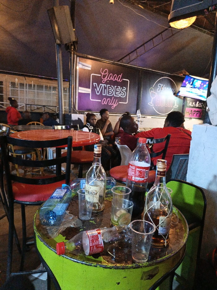 A crowded bar scene displays empty bottles and used cups on a vibrant green table. A red-topped table sits nearby with similar red chairs. Patrons are seated in the background, enjoying the ambiance. A sign reading "Good Vibes Only" is visible, along with another sign advertising "Happy Hour." A television screen flickers above the seated patrons. The setting appears to be dimly lit with casual, social energy.