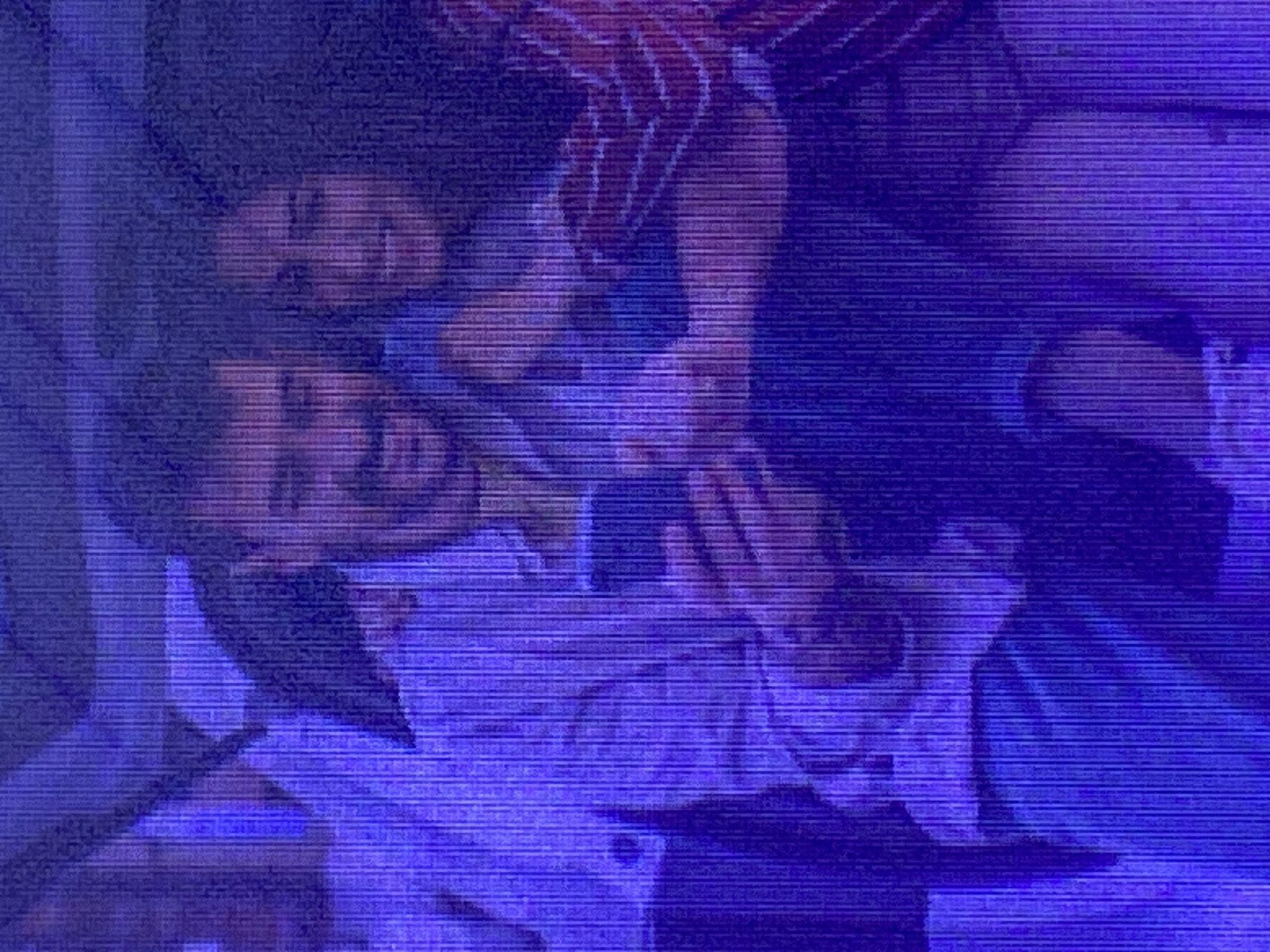 The image shows a close-up, low-resolution, and grainy view of two people, likely a couple, lying down and embracing. One person, with dark hair, is lying on their back, facing upwards. The other person, whose face is partially obscured by hair and is wearing a striped shirt, is leaning over them, with an arm wrapped around their neck. Their faces are close, suggesting intimacy or affection. The background is dark and indistinct, with a blueish tint, hinting at a dimly lit indoor setting, possibly at night. The overall impression is one of closeness and affection, with the subjects appearing relaxed and comfortable with each other. There is no visible text or discernible information about the time of day, weather, or specific location beyond the provided context of Ikorodu, Nigeria. The image quality makes it difficult to discern fine details about their clothing or physical features, but the posture and proximity of the subjects convey a sense of intimacy.