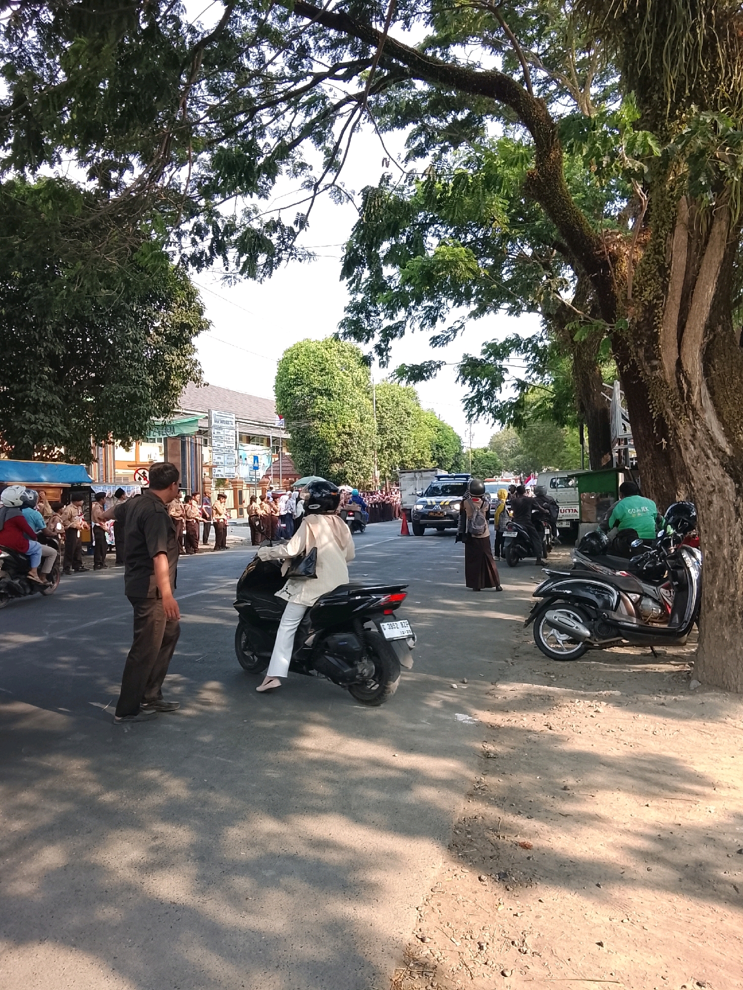 The image depicts a street scene in Slawi, Indonesia, likely in front of a school or official building, as indicated by the presence of uniformed students and a sign that appears to be from an Indonesian government or educational institution. A group of students in light brown uniforms and dark brown pants or skirts are standing in formation on the side of the road. Several people are on motorcycles, including a woman in a white outfit and helmet riding a black scooter, and a person in a green shirt with "GOJEK" written on it, also on a motorcycle. A man in a dark brown shirt and pants is standing in the foreground, seemingly directing traffic or observing the students. A police vehicle is visible in the background, parked on the left side of the road. The environment is characterized by mature trees with dense foliage casting shadows on the asphalt road and the sandy verge. The weather appears to be sunny and warm.