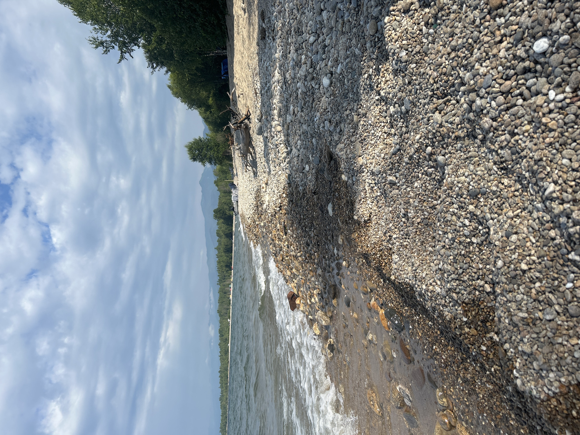 This image shows a rocky beach along a body of water, likely Lake Baikal, near Tolbazikha, Russia. The foreground is dominated by smooth, rounded pebbles in various shades of gray, brown, and white. Some of these rocks are wet from the lapping waves. The water, with its white foam and choppy surface, meets the shore at a gentle slope. Further inland, the beach transitions to sand, with a large piece of driftwood lying on it. In the background, a line of green trees forms a dense forest, beyond which mountains are visible under a cloudy sky. A few tents and what appear to be boats are sparsely scattered along the sandy shoreline, suggesting a recreational area. The overall atmosphere is tranquil and natural.