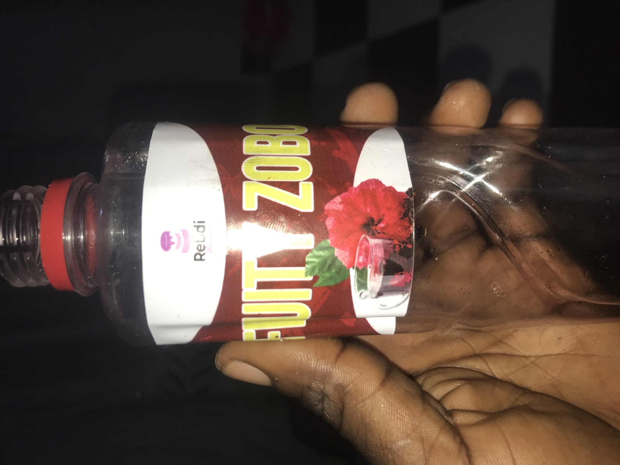 A close-up, horizontally oriented shot captures a dark-skinned hand holding a clear plastic bottle. The bottle features a red screw cap and contains a dark, reddish liquid, appearing partially consumed with air bubbles visible inside. A colorful label wraps around the bottle.
The most prominent text on the label, in large yellow letters against a maroon background, reads "FRUITY ZOBO". To the left of this main text, a smaller white section of the label displays a purple cupcake-like logo and the partial word "ReLdi". To the right of the "FRUITY ZOBO" text, a vivid red hibiscus flower is depicted, accompanied by a clear glass filled with a red beverage.
The background is predominantly dark, suggesting an indoor setting with dim lighting, possibly evening or night. A subtle black and white checkered pattern is faintly visible in the upper right corner of the background. The hand holds the bottle casually with fingers wrapped around it.
This media file is from Rafin Par, Nigeria.