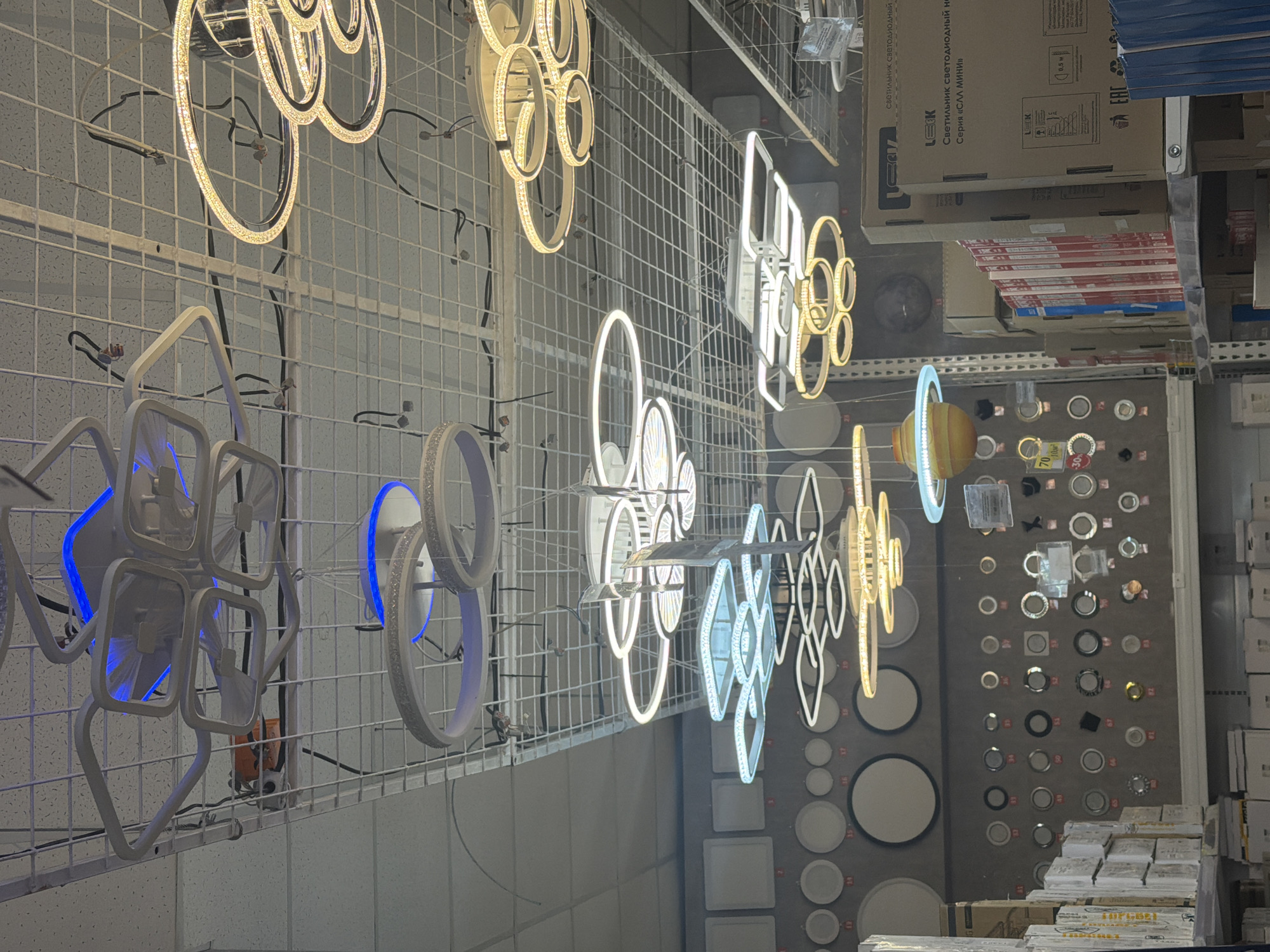 This is an indoor shot in what appears to be a store or warehouse setting, likely a lighting department, possibly in Chaikovsky, Russia. Various modern light fixtures and lamps are displayed on wire shelving. The lighting fixtures are in different designs and shapes, many with integrated LED lighting in circular, square, and abstract shapes. Some of the fixtures are illuminated with white or blue LED light.
Boxes and packaging are stacked on the shelves, presumably containing the merchandise. A display board showcases different types of lights, including recessed lighting and spotlights. The overall environment appears to be a retail or storage space with a focus on lighting products. No people are visible in the frame.