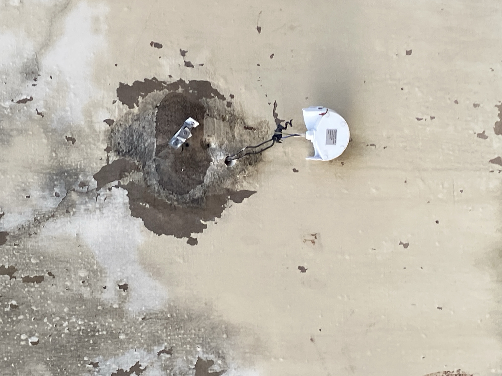 The image depicts a damaged wall with a circular hole from which wires and a detached white light fixture hang. The wall exhibits peeling paint and discoloration, suggesting neglect or damage. A small metallic object is lodged in the wall near the hole. The scene is devoid of any people or discernible activity. The context provided places this image in Jauro Yinu, Nigeria, an area known for its unique geographical and architectural features, though specific details about the immediate surroundings are not visible in the image.
