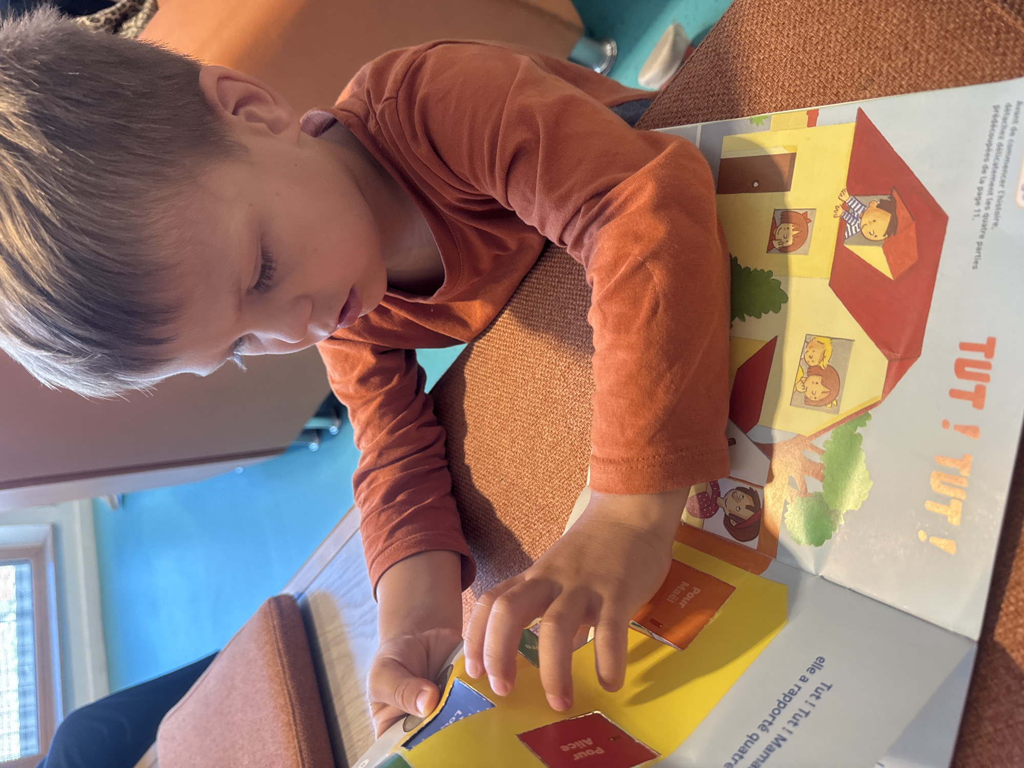 A young Caucasian boy with short, light brown hair is sitting at a wooden table, intently looking down at a colorful pop-up book. He is wearing an orange long-sleeved shirt. His hands are placed on the open pages of the book, with his fingers touching a yellow, angled flap. The book features bright illustrations, including depictions of children in windows and houses. Text in French is visible on the pages, with some words appearing upside down. The table's surface is covered with a textured, orange fabric. In the background, a blue floor and part of a chair are visible. The overall lighting suggests an indoor setting, possibly a library or classroom, given the presence of what appears to be a window with a view of trees. This scene is set in Saint-Claude, France.