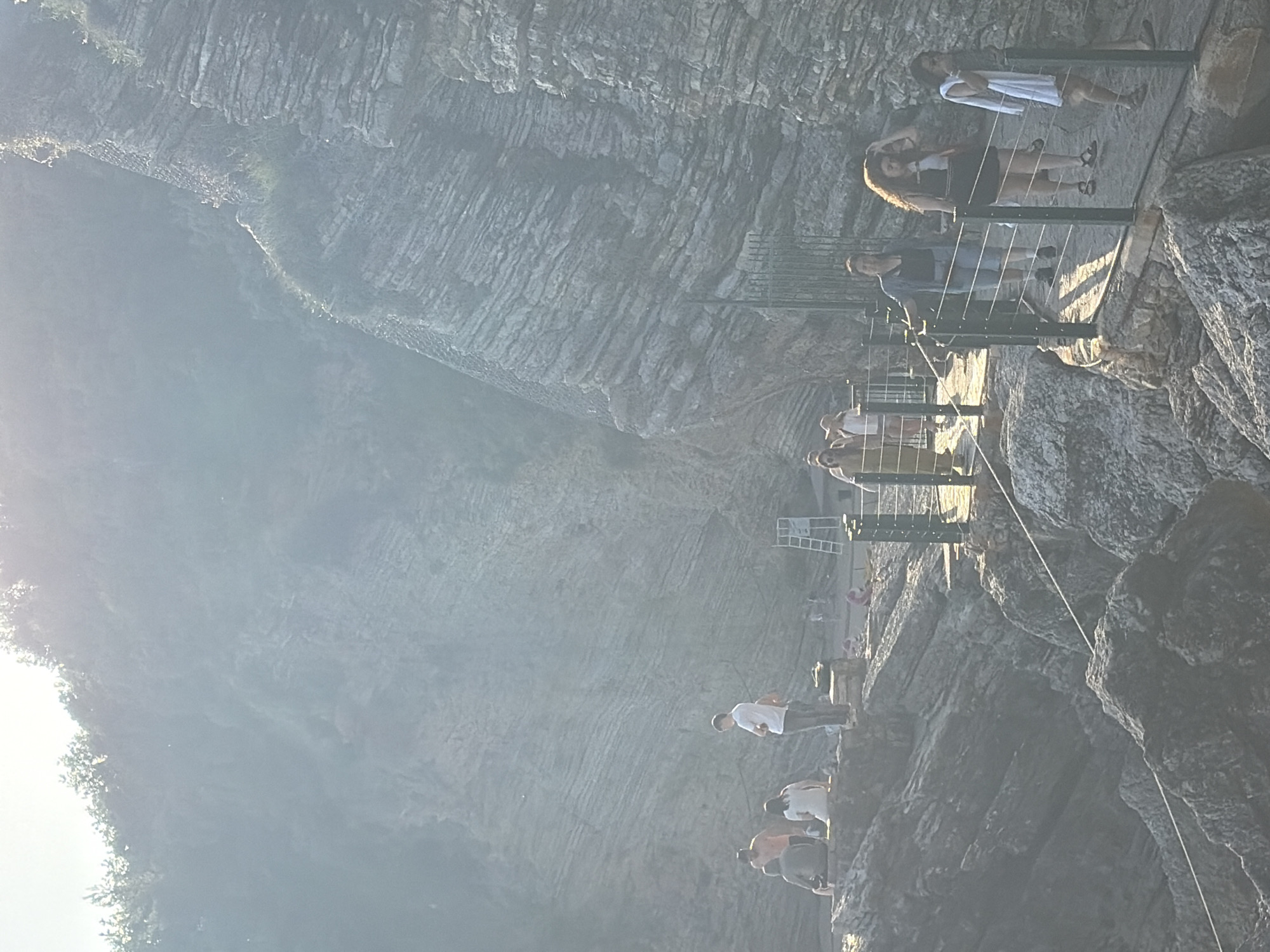 The image depicts a group of people walking on a narrow, elevated pathway built along a steep rock face. The pathway is secured with metal railings. The environment consists of a rocky cliff or mountain with visible geological layering. The setting is partially obscured by bright sunlight or fog. It looks like it is at the city of Budva, Montenegro, along a narrow elevated pathway made up of metal rails with a group of people.