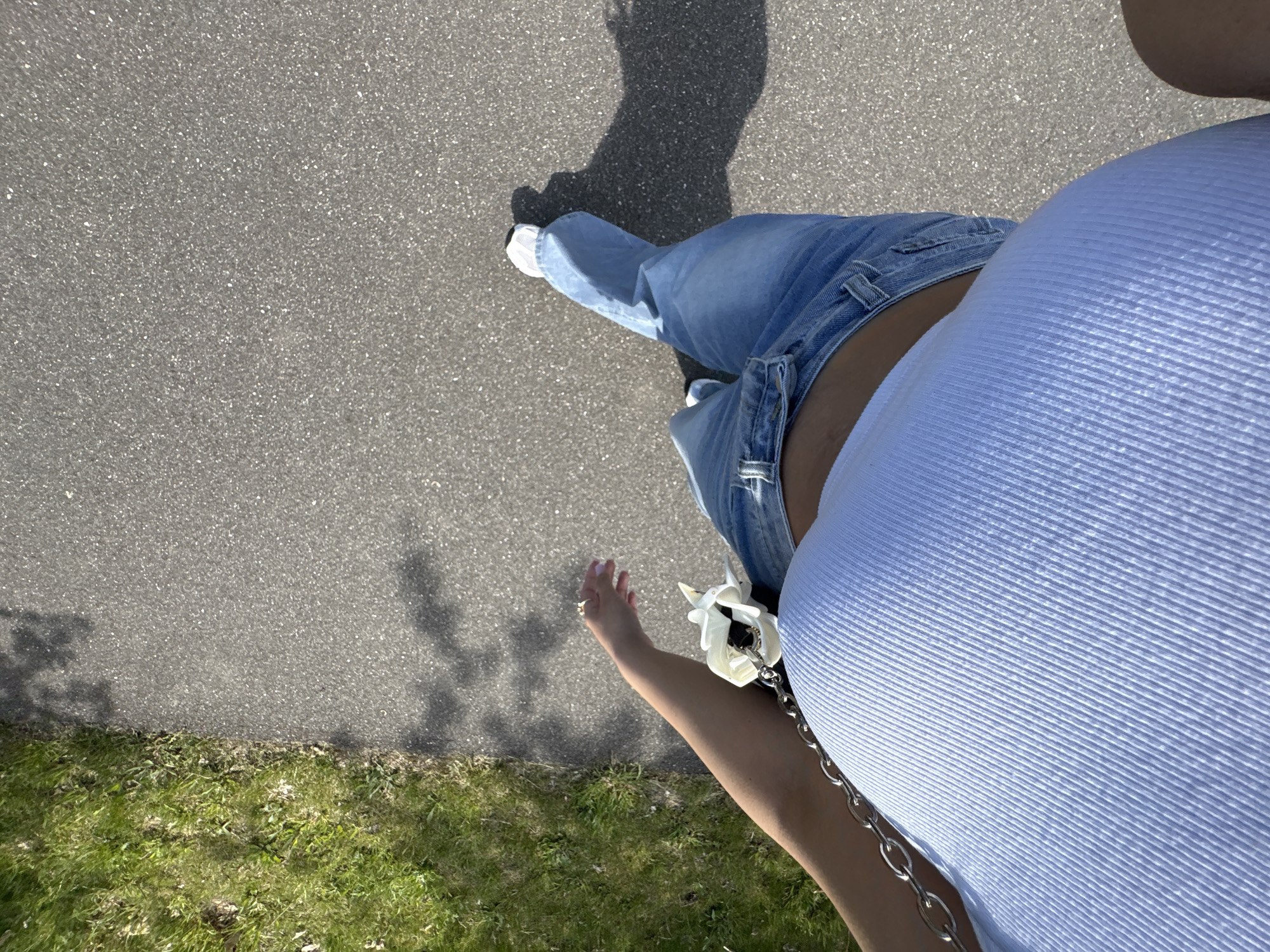 The image shows a first-person perspective of a person walking on an asphalt surface, potentially a sidewalk or path. The person is wearing blue jeans, white sneakers, and a light blue tank top with a silver chain. A shadow of the person is visible on the asphalt. The edge of a grassy area is seen on the left side of the image. The location is in Wassenberg, Germany.