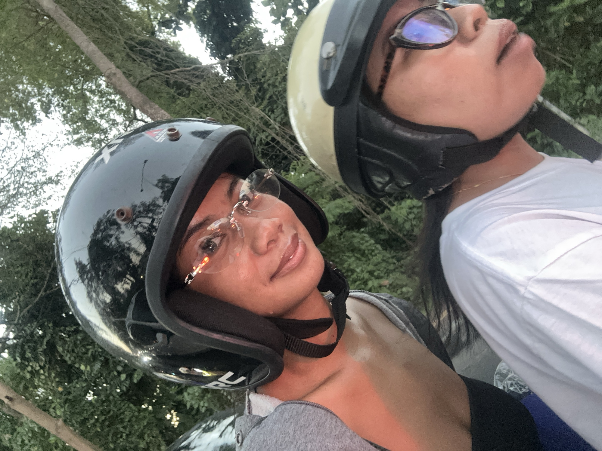 Two individuals, likely female based on their features, are positioned on a scooter, wearing helmets. The person in the foreground, on the right, wears a light yellow helmet and eyeglasses with dark frames, and a white t-shirt. The person in the background, on the left, wears a black helmet, eyeglasses with thin, decorative frames, and a grey hooded jacket over a dark top. They appear to be riding during daylight, with trees and foliage visible in the background, suggesting a rural or park-like setting within or near Denpasar, Indonesia. The lighting indicates it might be late afternoon or early evening.